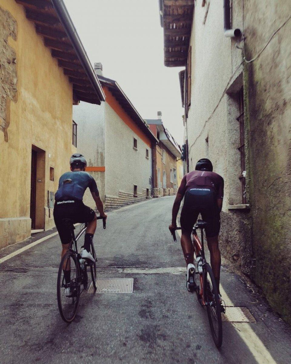 Two cyclists in Mechanism jerseys riding through narrow European village street