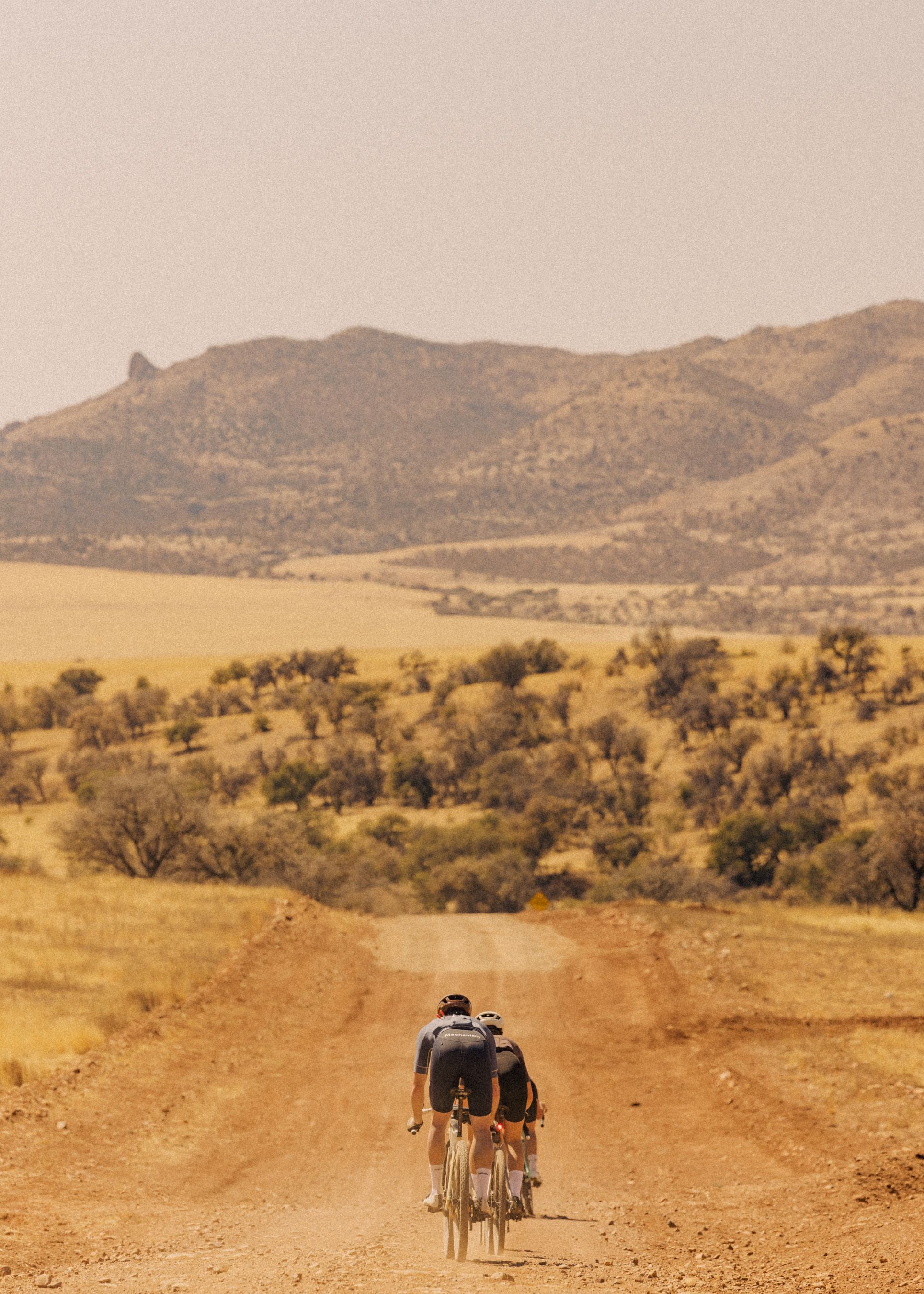Two cyclists riding on desert dirt road with mountains in background