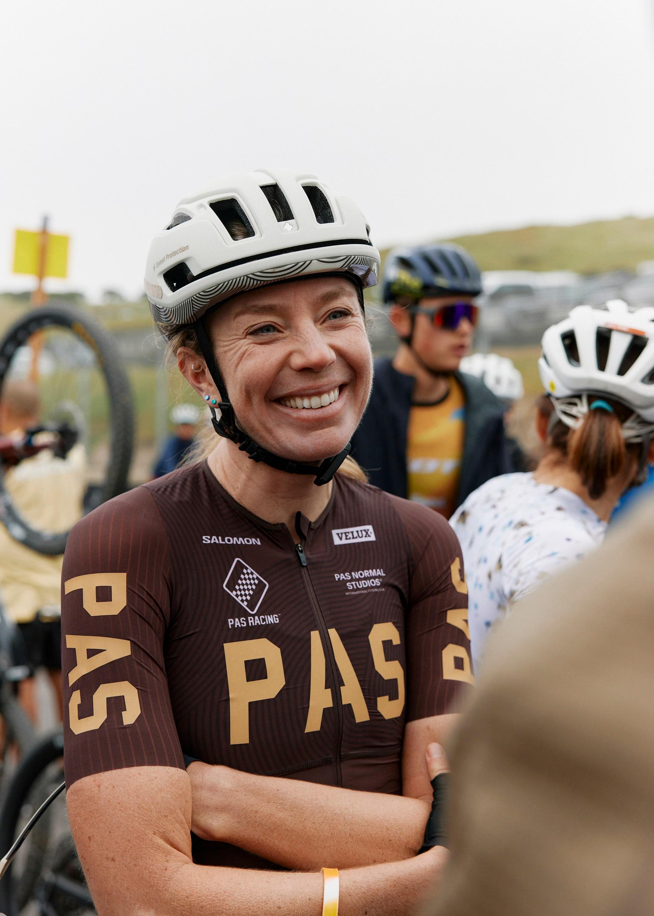 Female cyclist wearing Women's PAS brown racing jersey with gold lettering and white helmet, smiling at camera