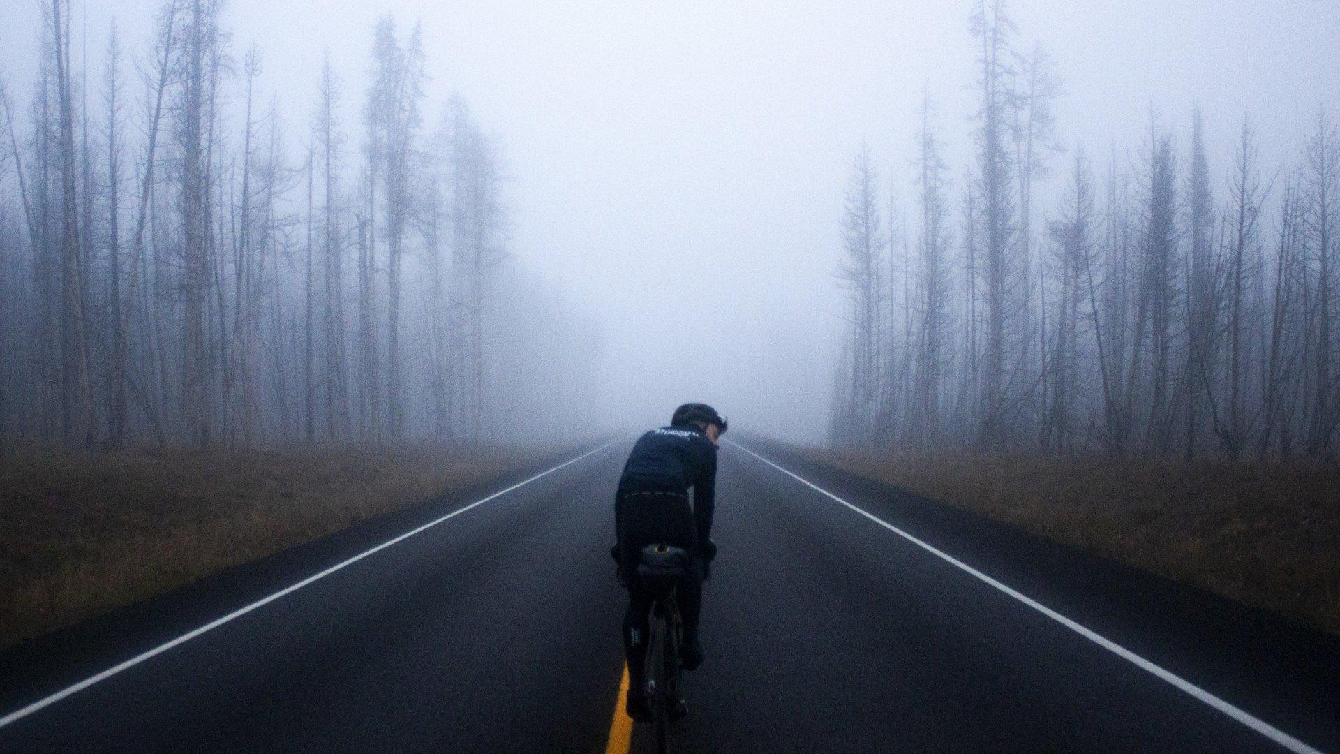 Cyclist in black apparel riding through misty forest road with bare trees