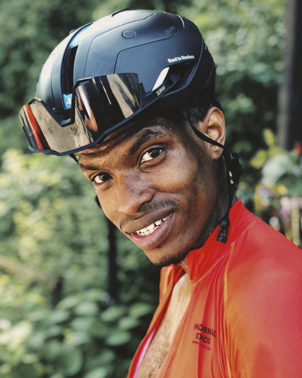 Model wearing orange cycling jersey and black helmet with reflective visor, smiling outdoors