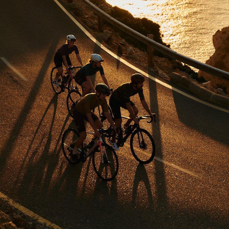 Four cyclists riding on coastal road at sunset with ocean backdrop and dramatic shadows