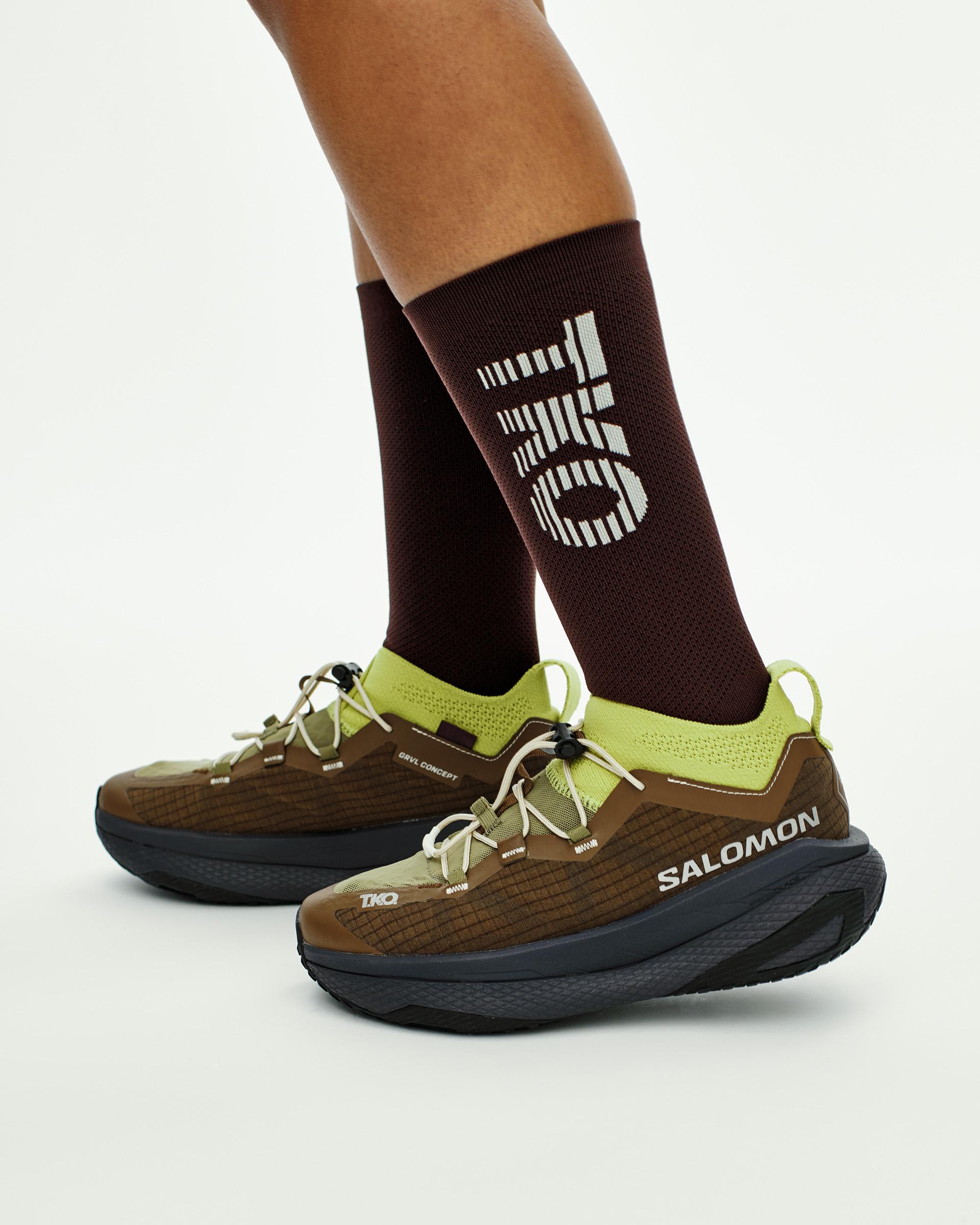 Green Salomon T.K.O. GRVL CONCEPT trail running shoes on model with brown socks featuring white logo