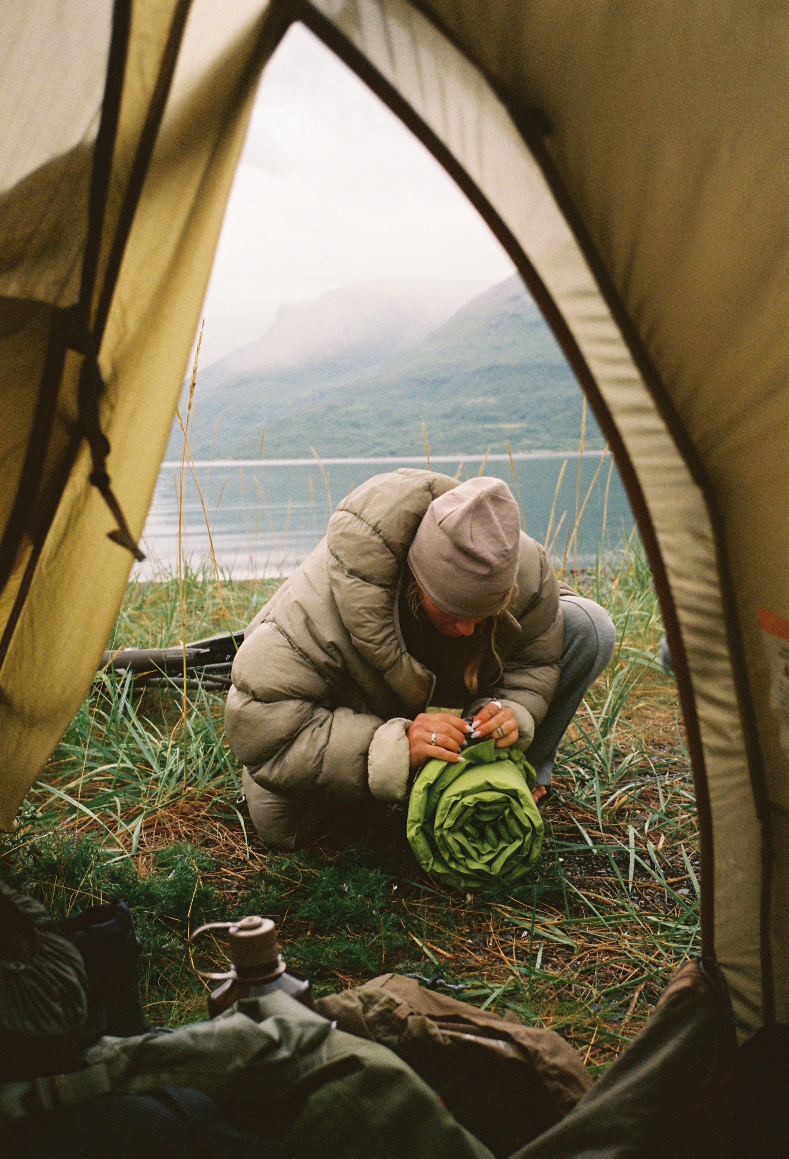 Model wearing beige down jacket and green base layer camping by coastline, view from inside tent