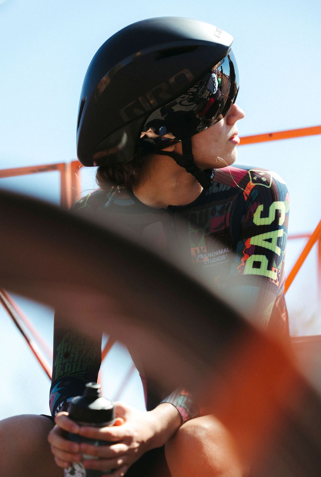Model wearing dark aerodynamic helmet and colorful cycling jersey with green PAS lettering in sunny outdoor setting
