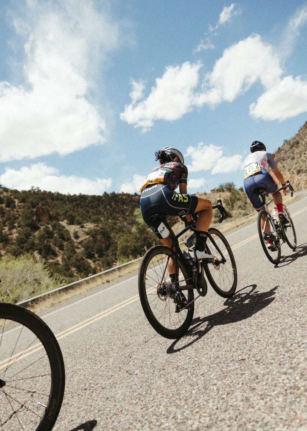 Two cyclists in aerodynamic position riding time trial bikes on desert road with mountainous landscape