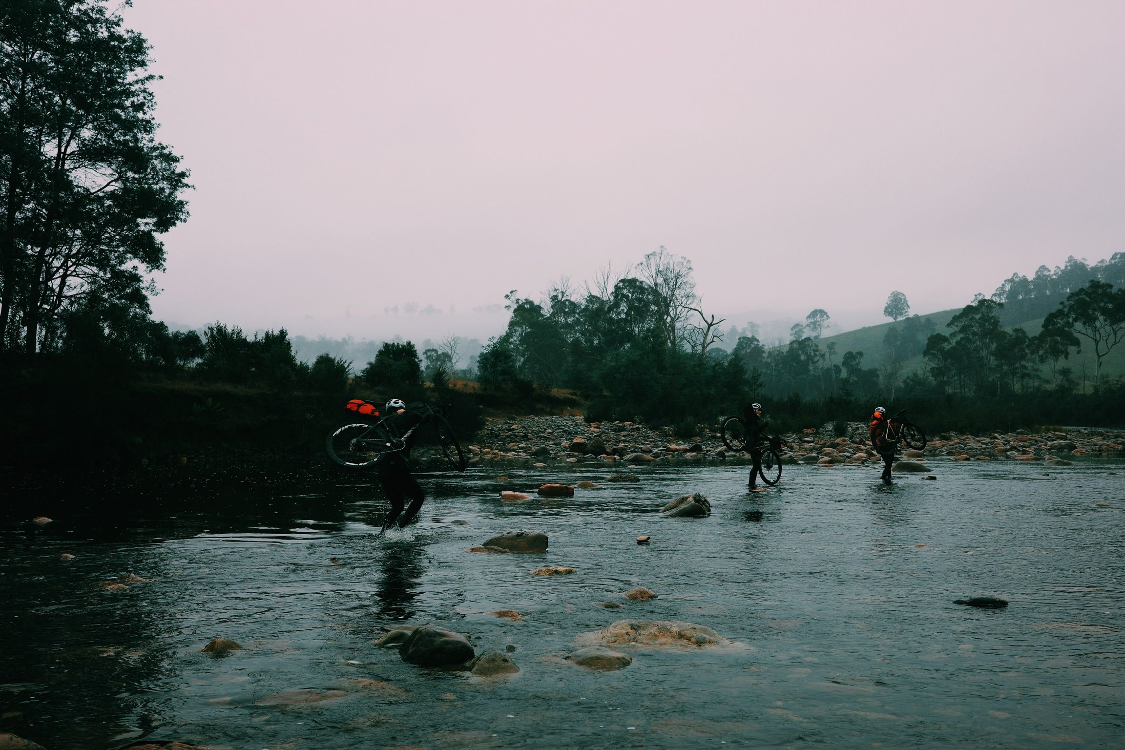 Cyclists crossing rocky river with bikes during dawn in Tasmanian wilderness