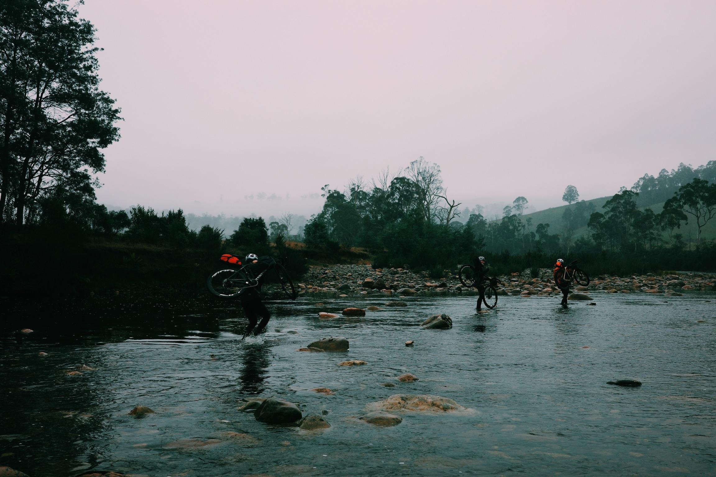 Cyclists crossing rocky river with bikes during dawn in Tasmanian wilderness