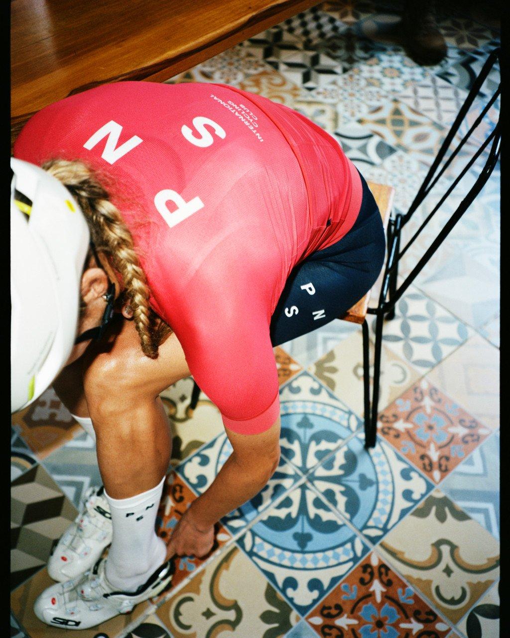 Model wearing pink long-sleeve jersey with collection branding stretching on patterned tile floor