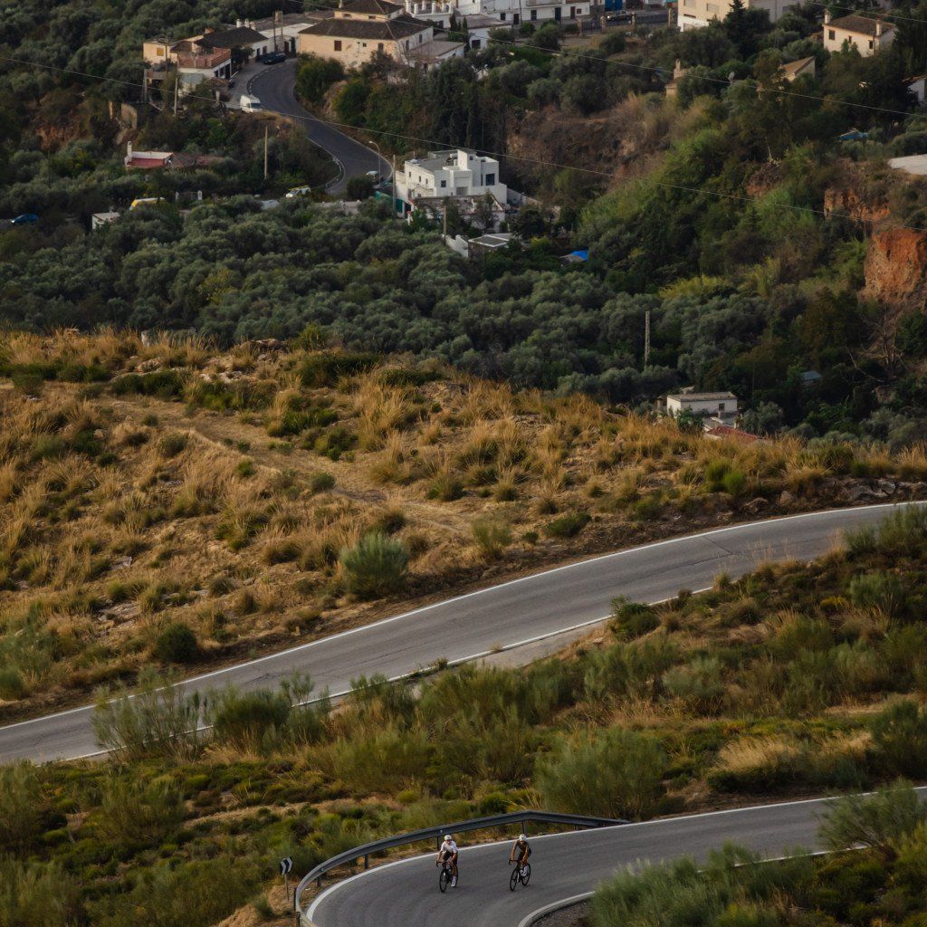 Two cyclists riding on winding mountain roads through Mediterranean hillside village at golden hour