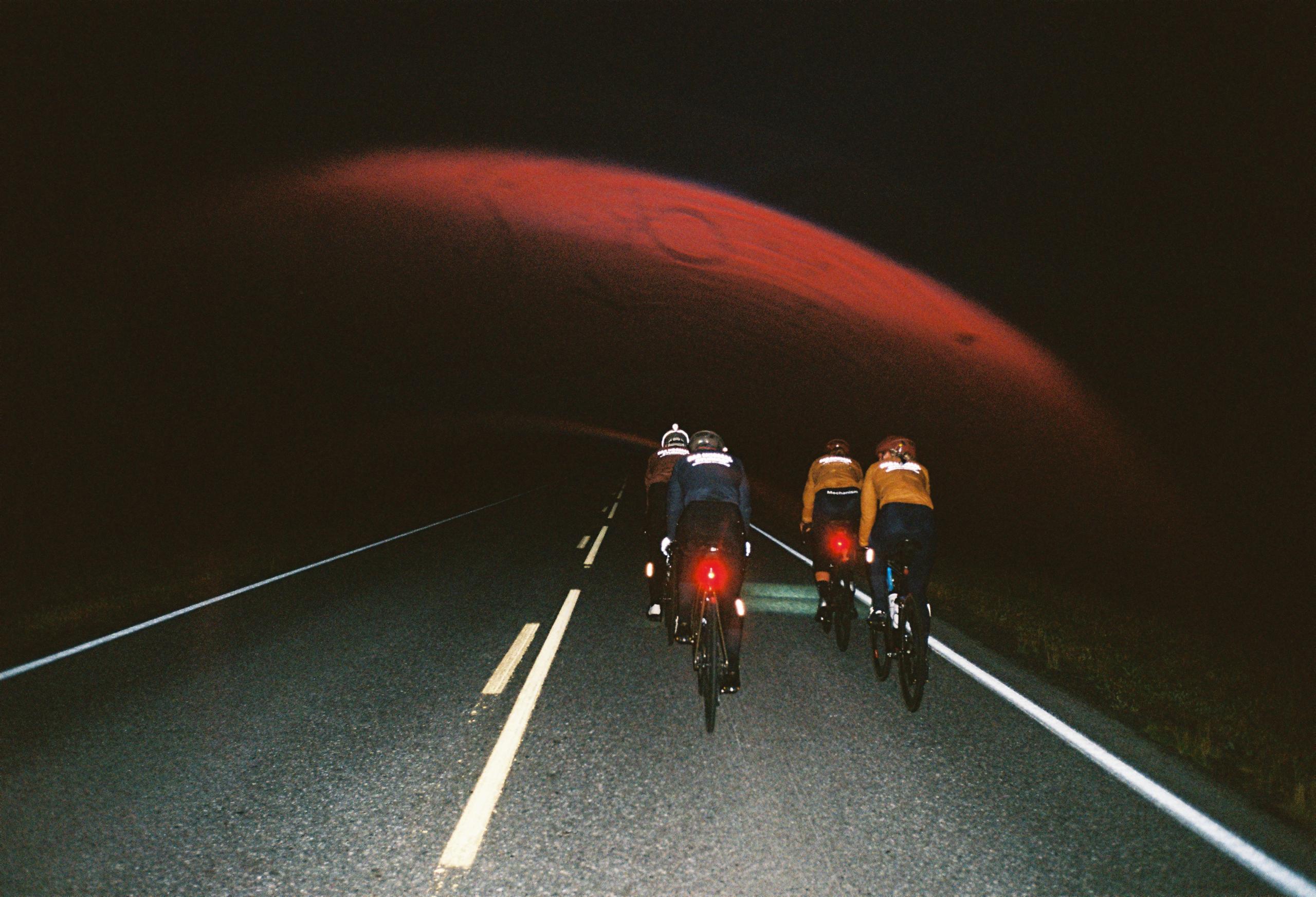 Two cyclists riding on dark road with red lights under dramatic red sky at dusk