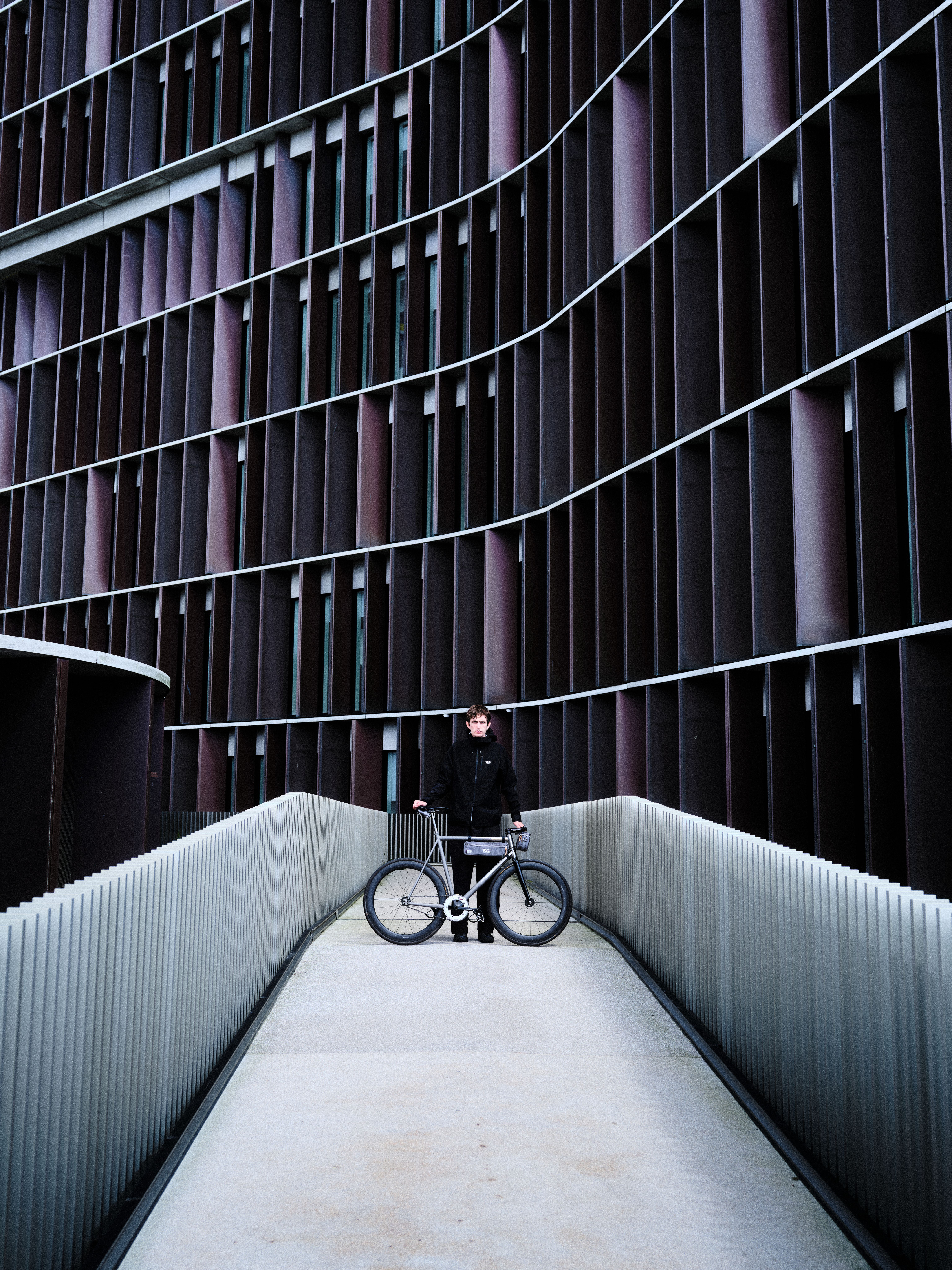 Model wearing black cycling apparel with bicycle on modern concrete walkway against geometric building facade