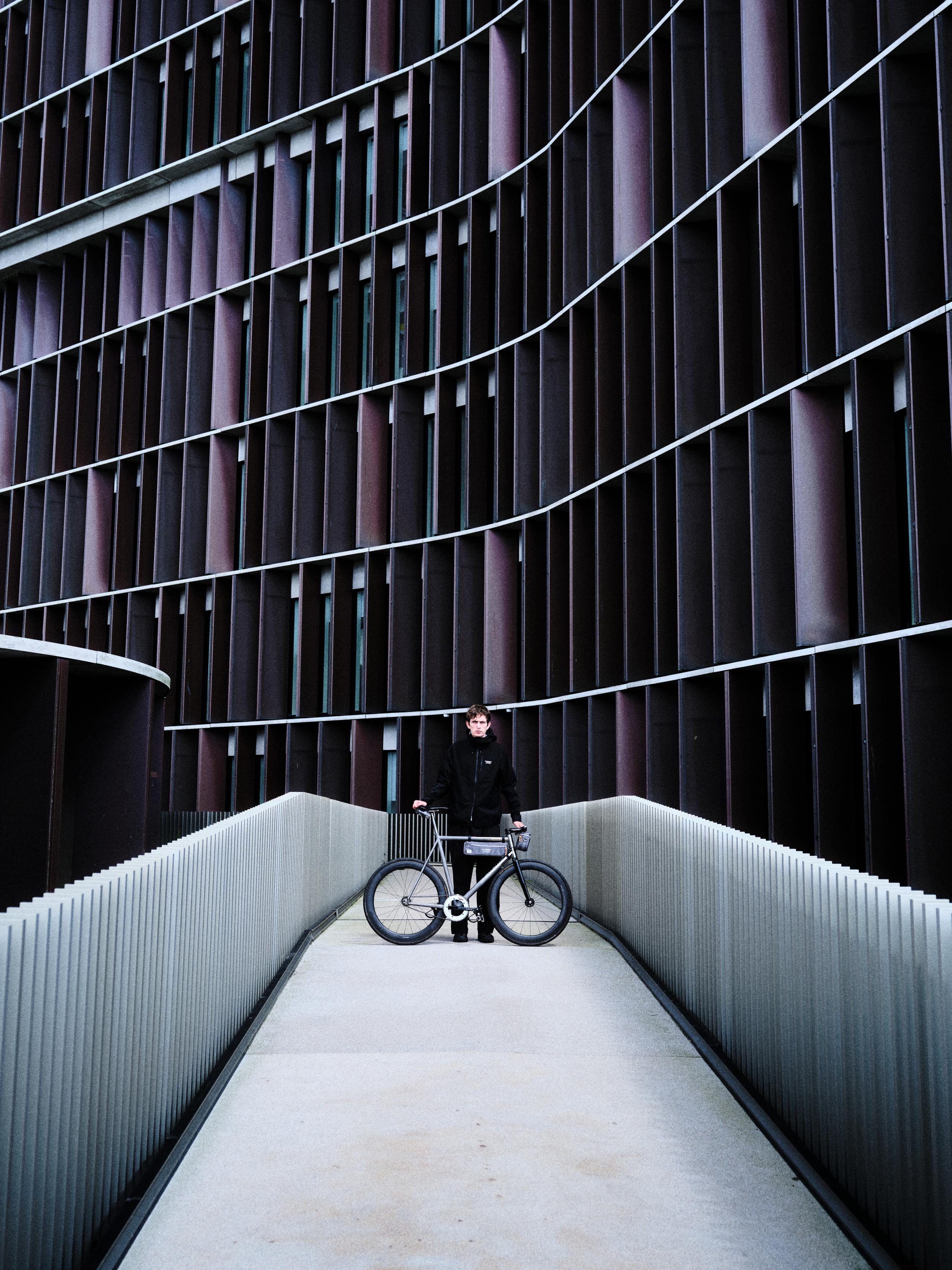 Model wearing black cycling apparel with bicycle on modern concrete walkway against geometric building facade