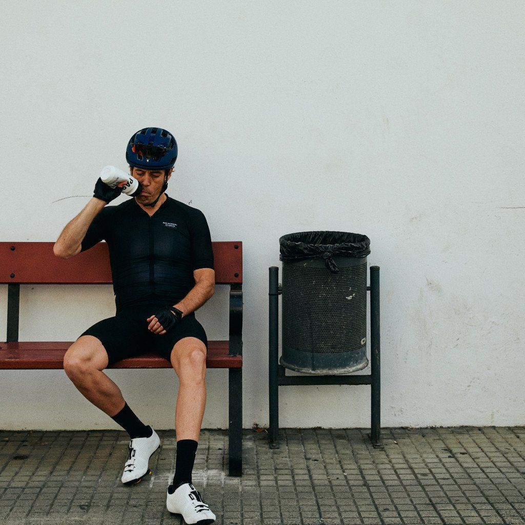 Model wearing black cycling jersey and shorts drinking from bottle, seated on bench with blue helmet