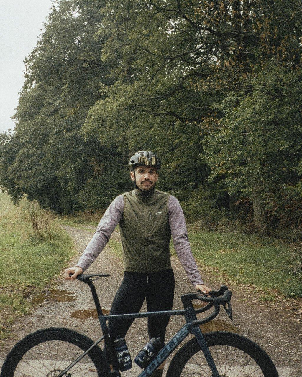 Model wearing olive green vest with grey long sleeves standing with road bike on country path