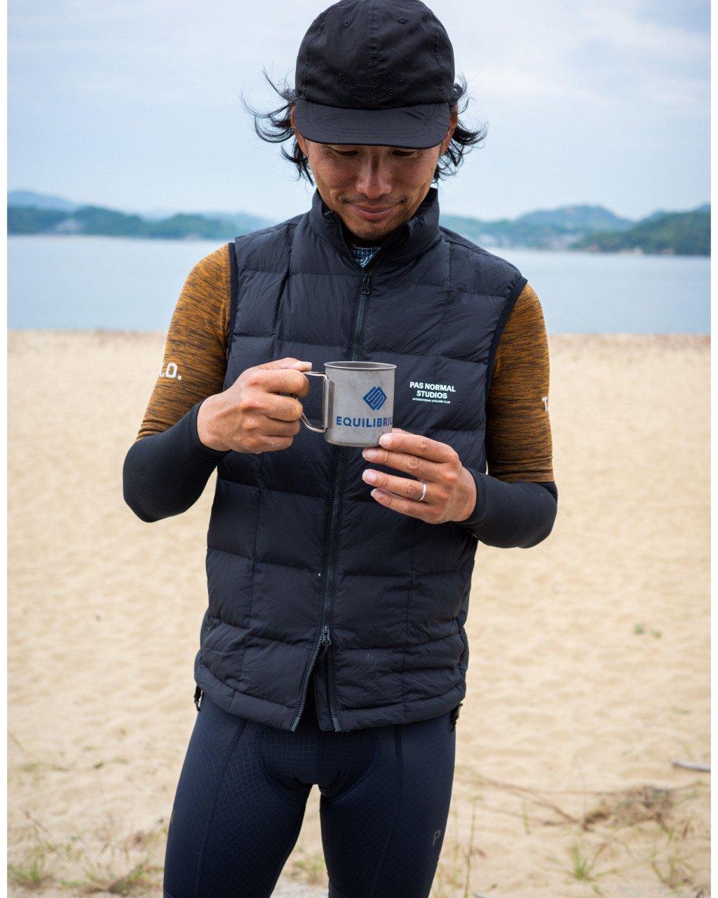 Model wearing black quilted vest with logo, brown long sleeve jersey, black cap, holding mug on beach