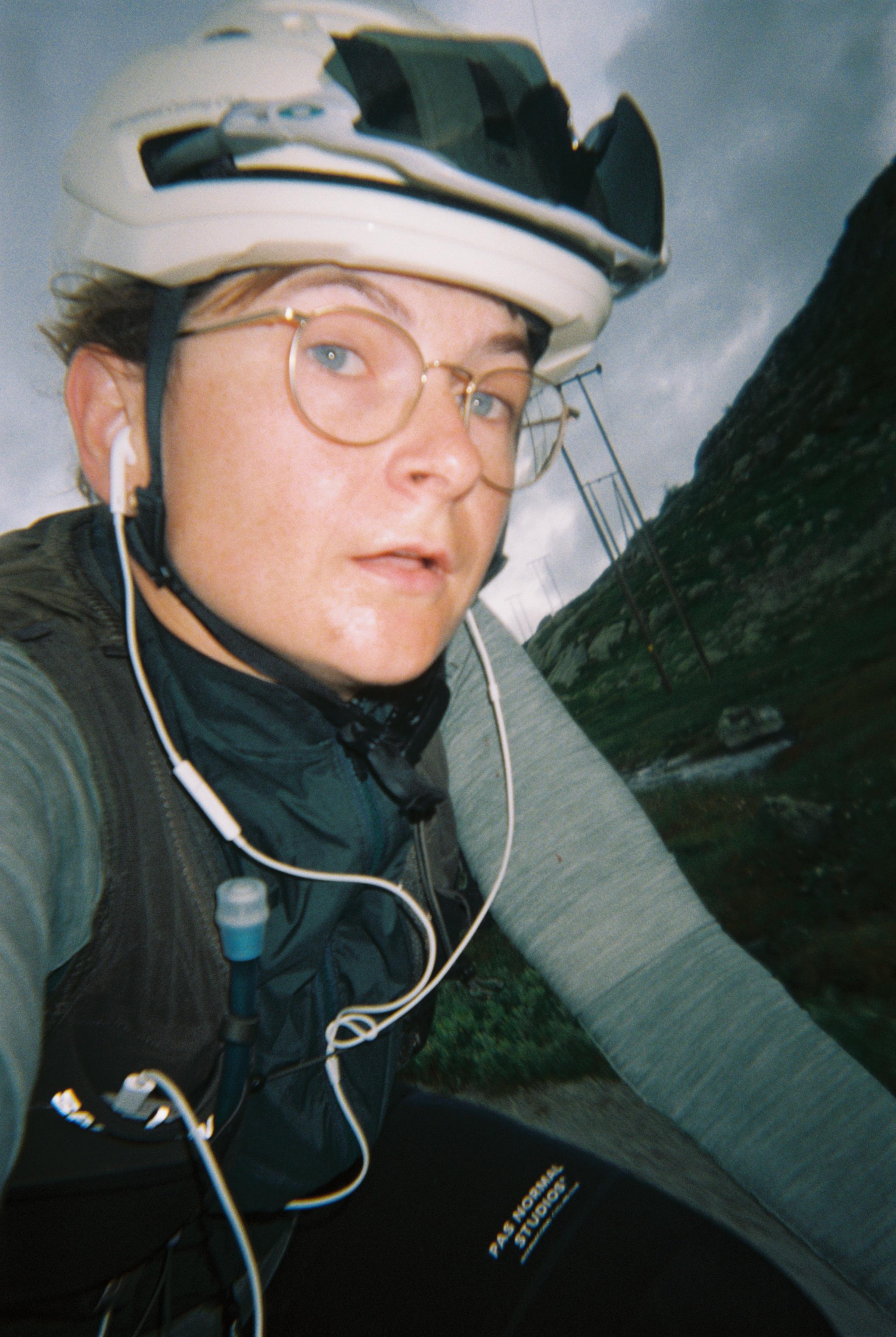 Model wearing white cycling helmet with earbuds, glasses, and dark cycling clothing in mountainous setting