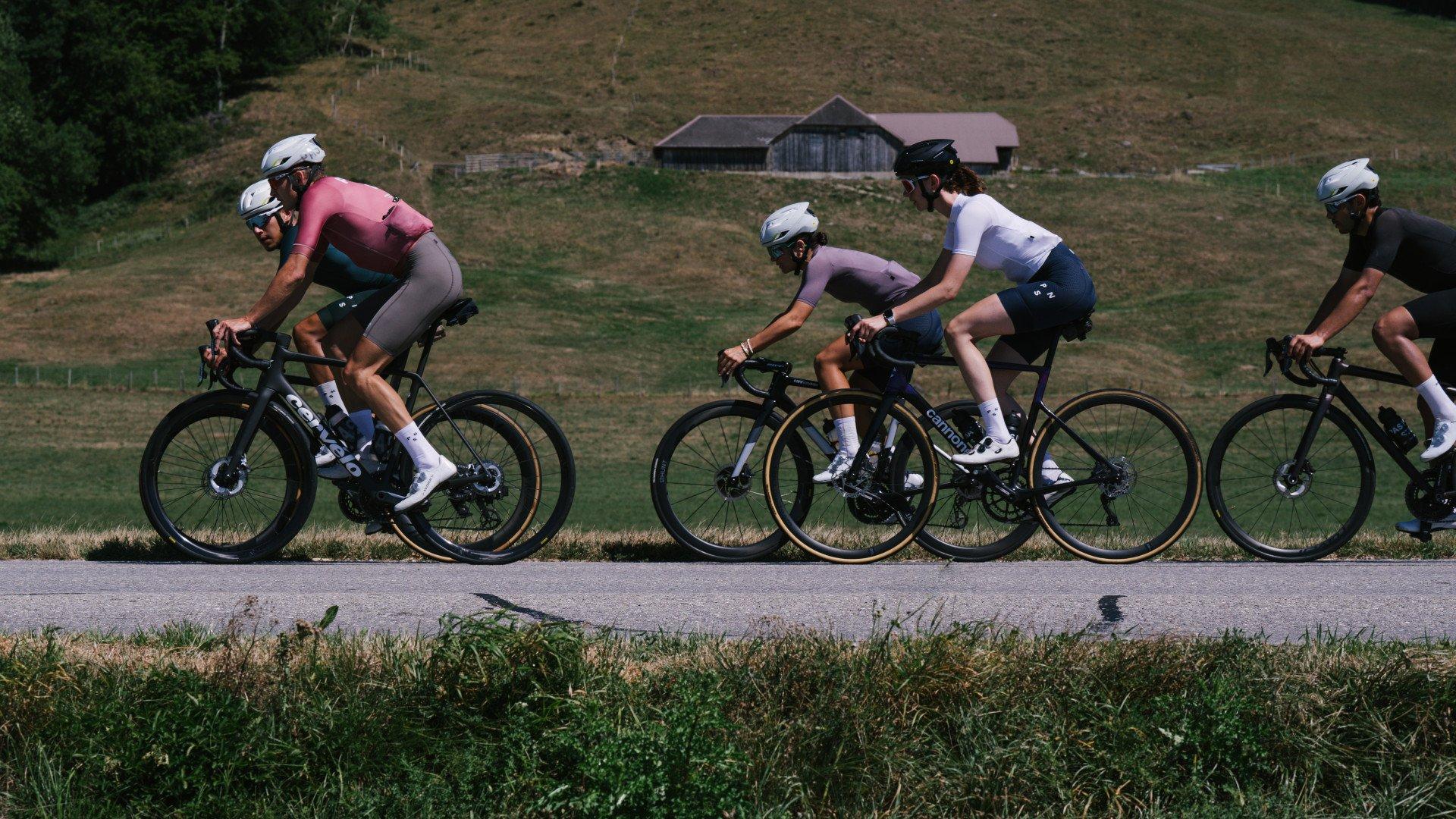 Four cyclists in aerodynamic position riding time trial bikes on rural road wearing colorful cycling kits