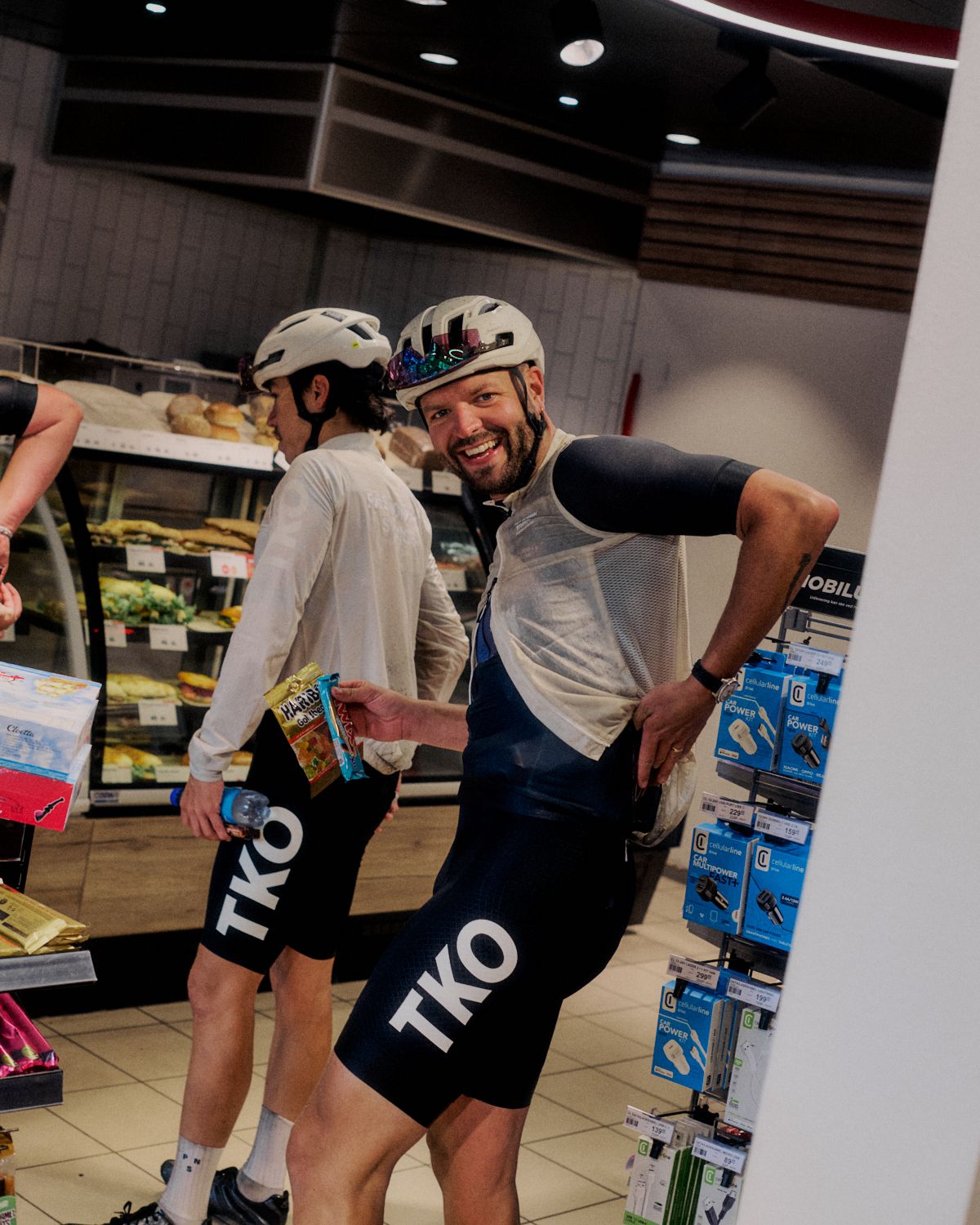 Two cyclists in helmets shopping in store, one wearing grey jersey and black bib shorts with logo branding
