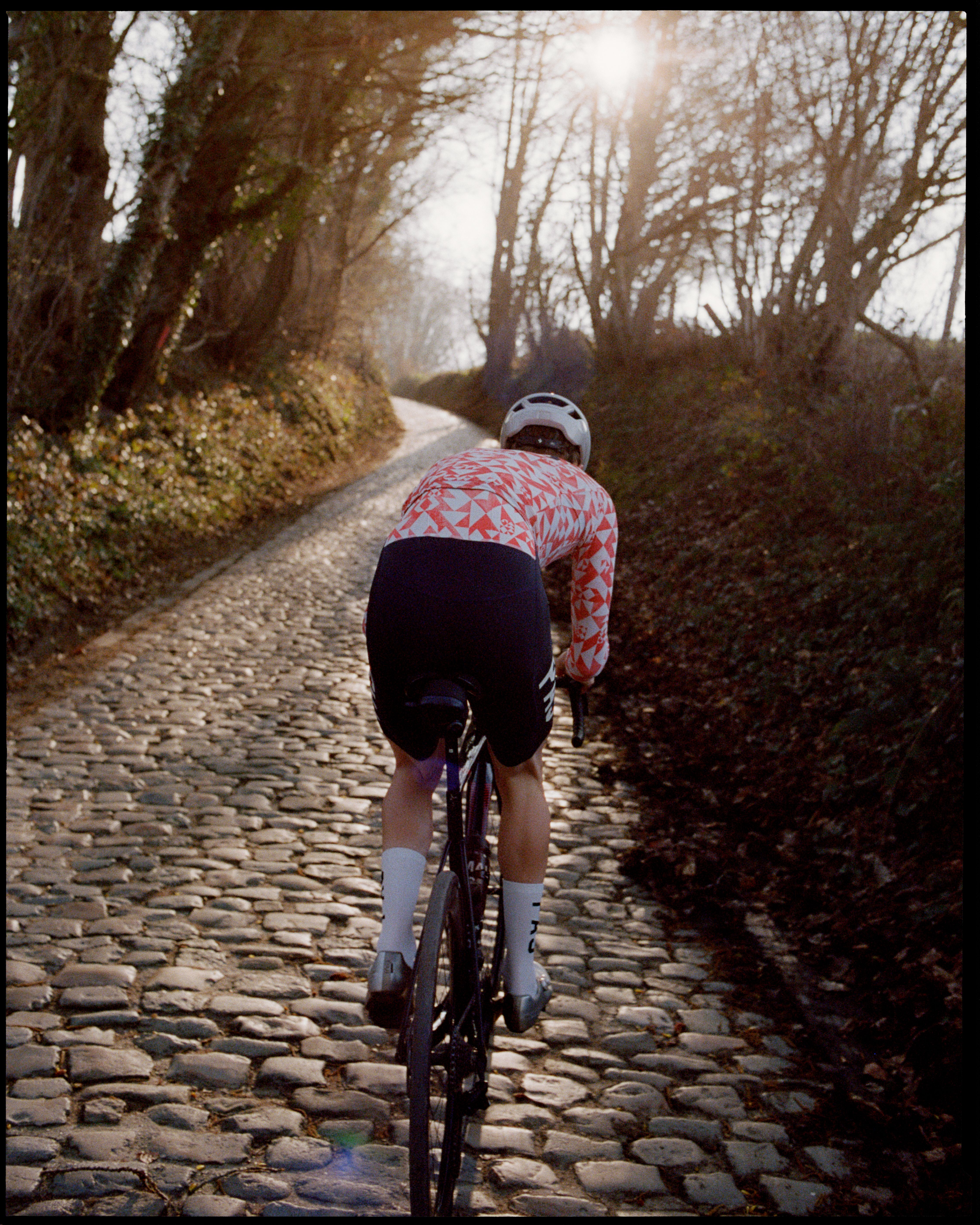 Model wearing red and white geometric patterned jersey with black shorts cycling on cobblestone path through trees