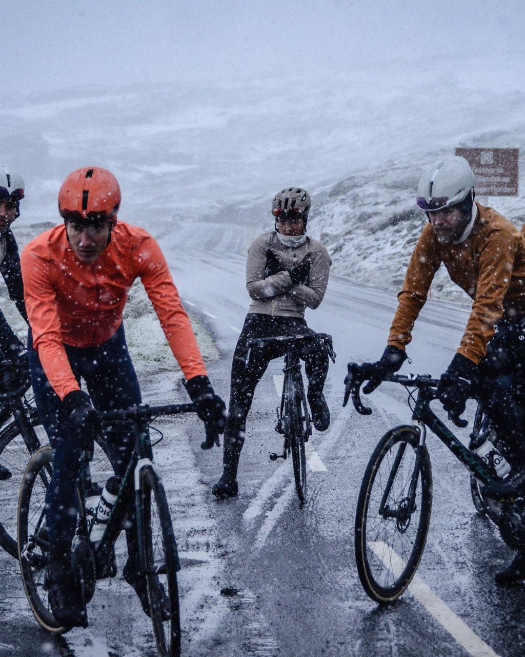 Cyclists in orange, brown and grey jerseys riding through heavy snowfall in snowy mountains