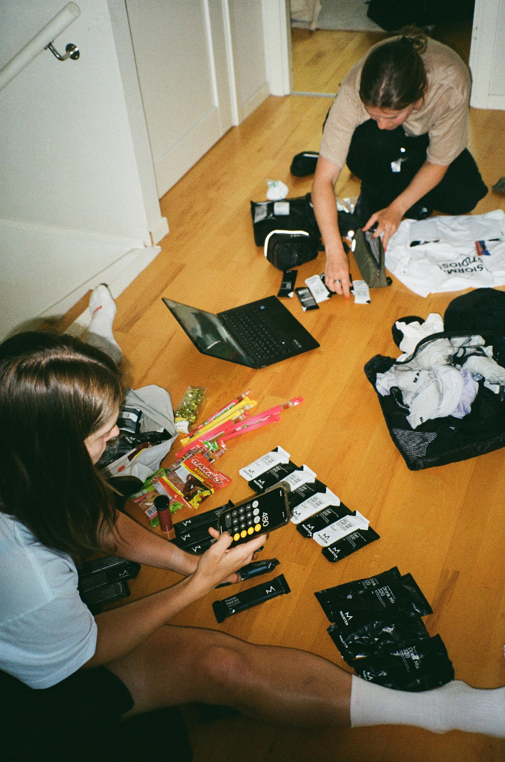 Two people organizing cycling apparel packages on wooden floor with laptop and snacks