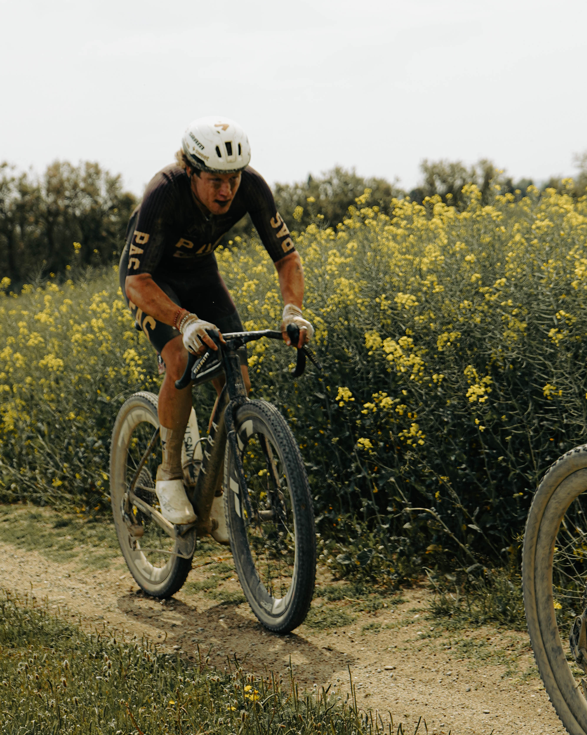 Model wearing black PAS Racing skinsuit cycling on gravel path through yellow wildflower field