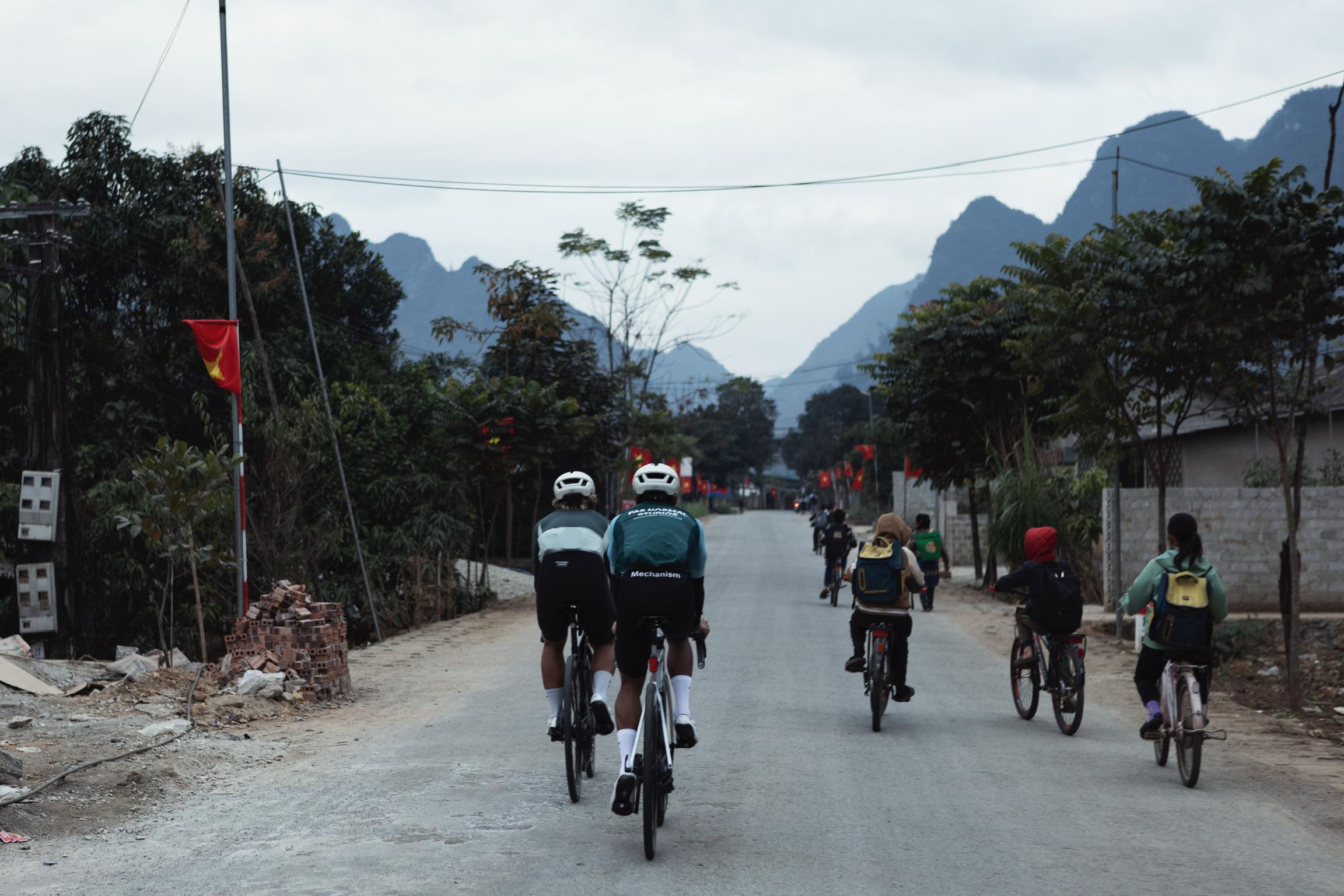 Two cyclists wearing Mechanism jerseys riding on rural road with mountain backdrop and local residents