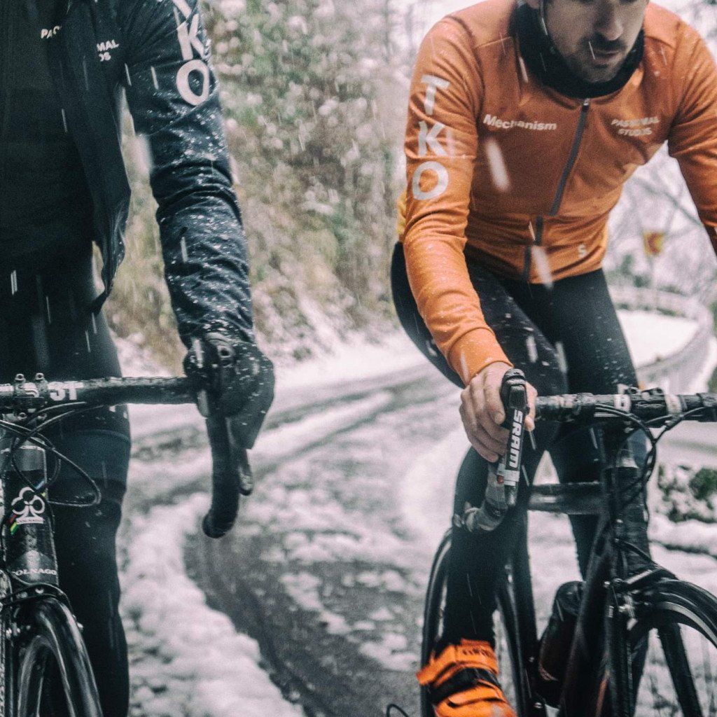 Models wearing orange Mechanism jerseys and black tights cycling on gravel road