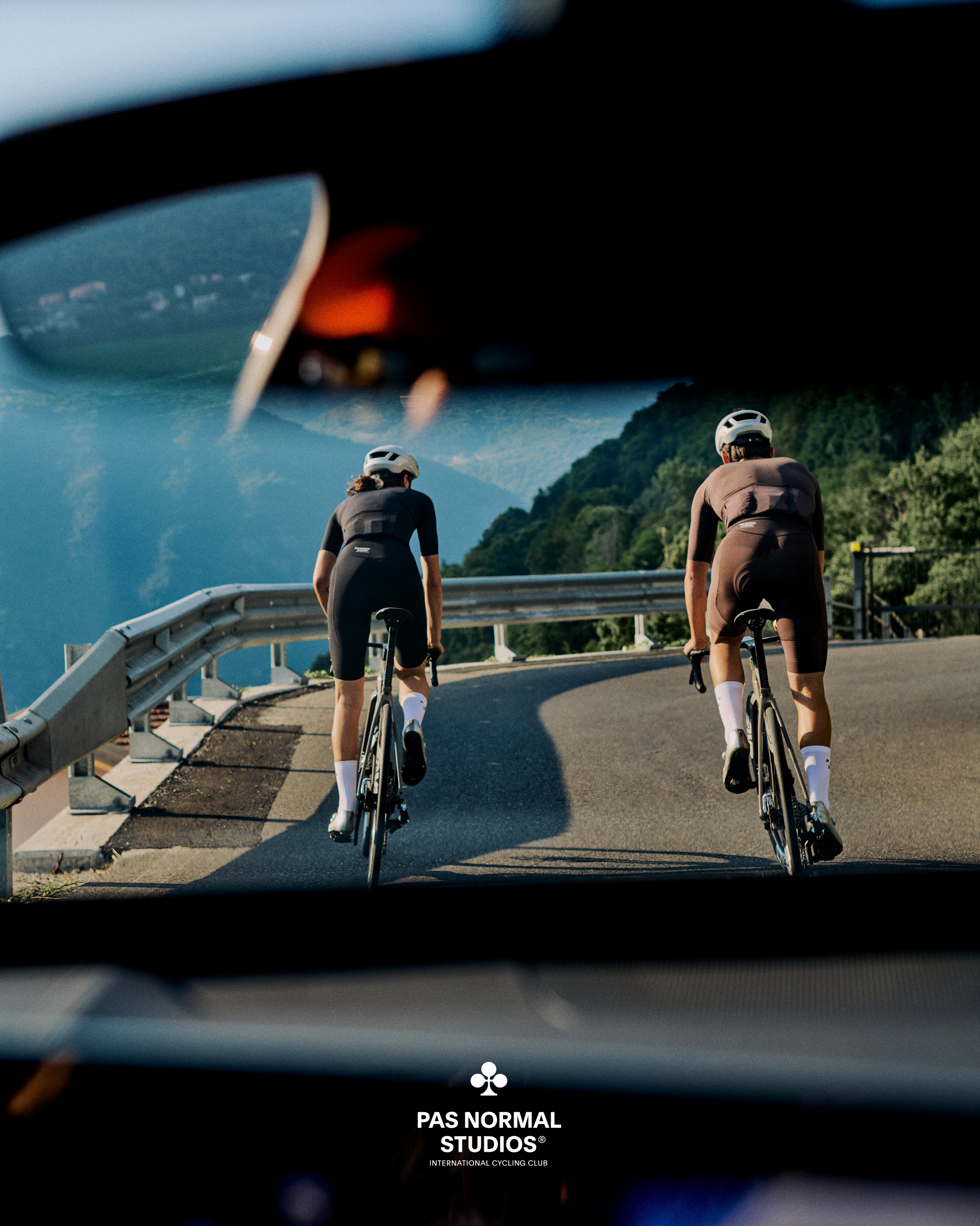 Two cyclists in aerodynamic position on time trial bikes crossing bridge, viewed through car windshield