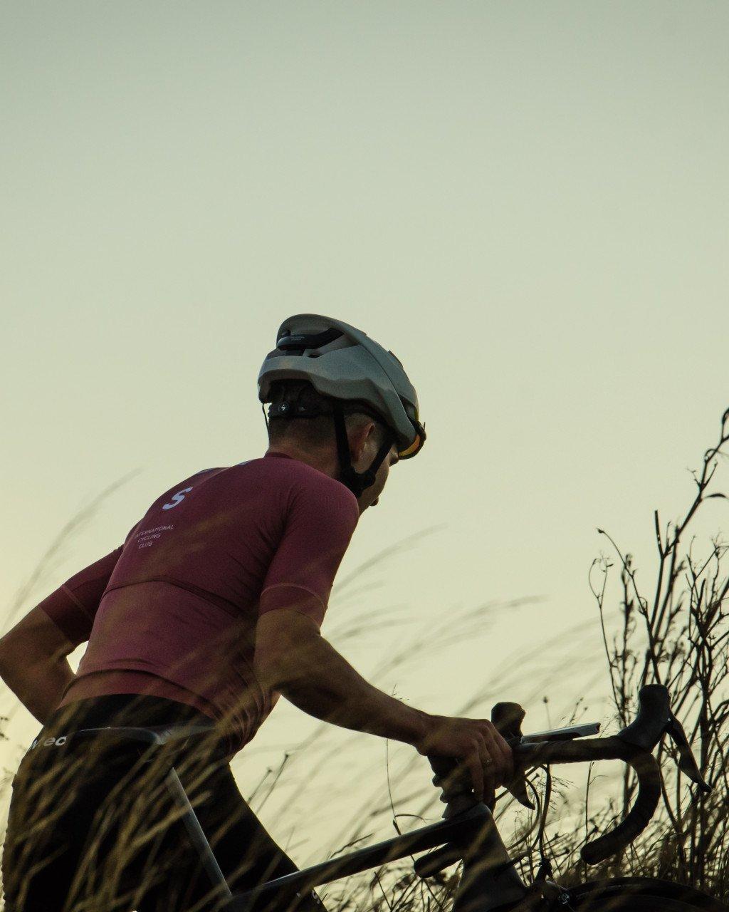 Model wearing burgundy cycling jersey with chest branding, cycling outdoors during golden hour