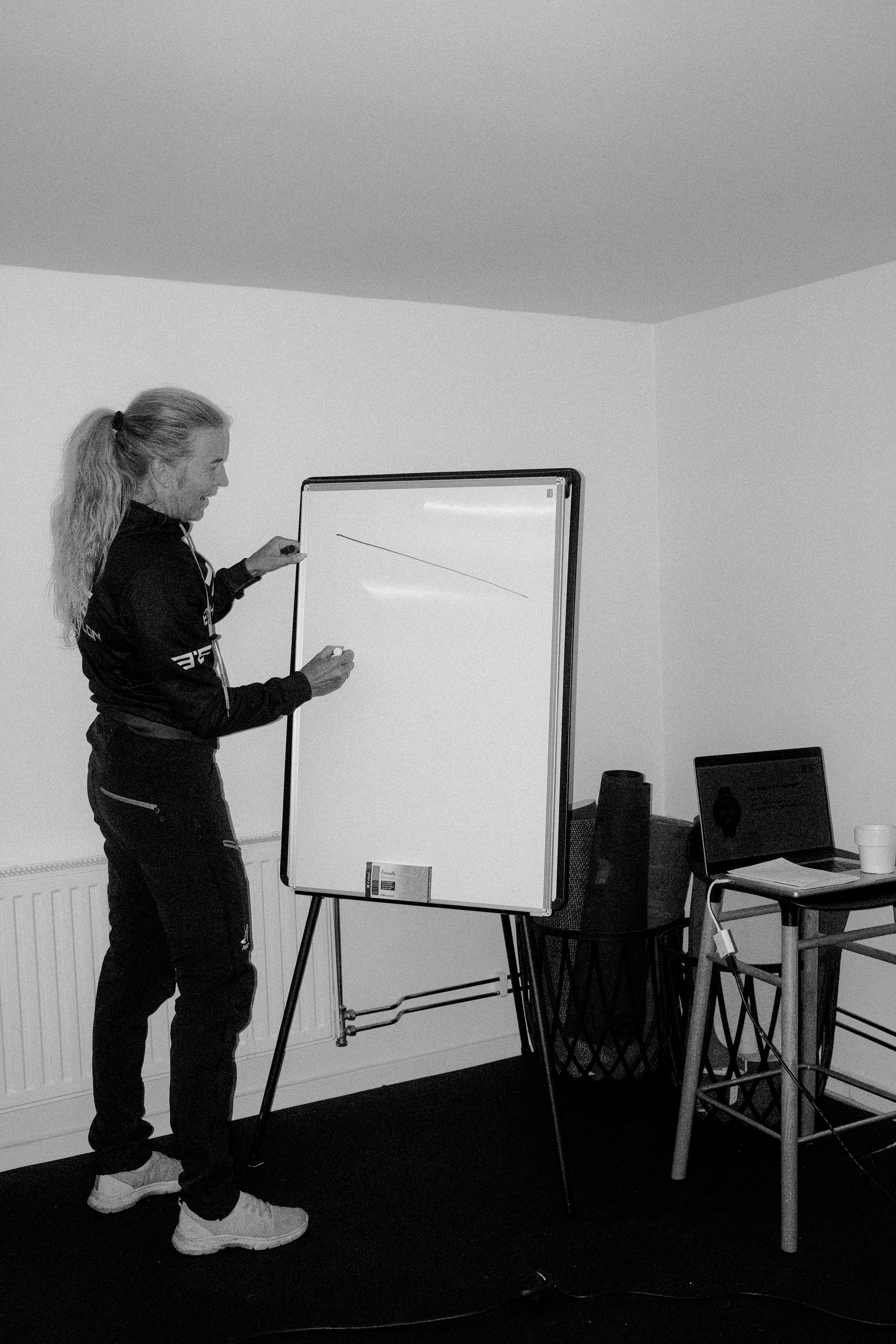 Model wearing black cycling kit with logo branding presenting at whiteboard in office setting