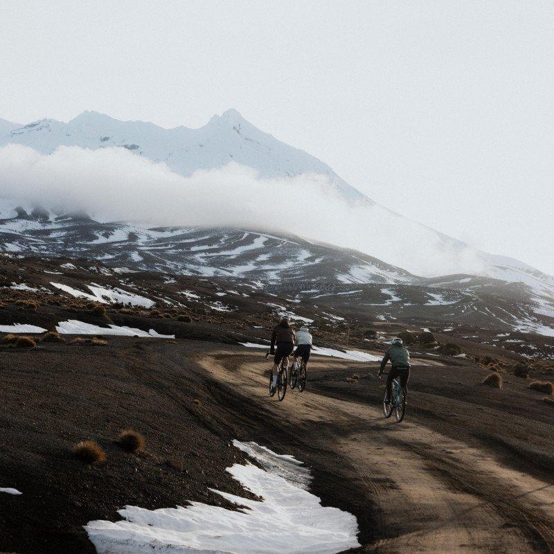 Cyclists riding gravel road through snow-covered volcanic landscape with misty mountains