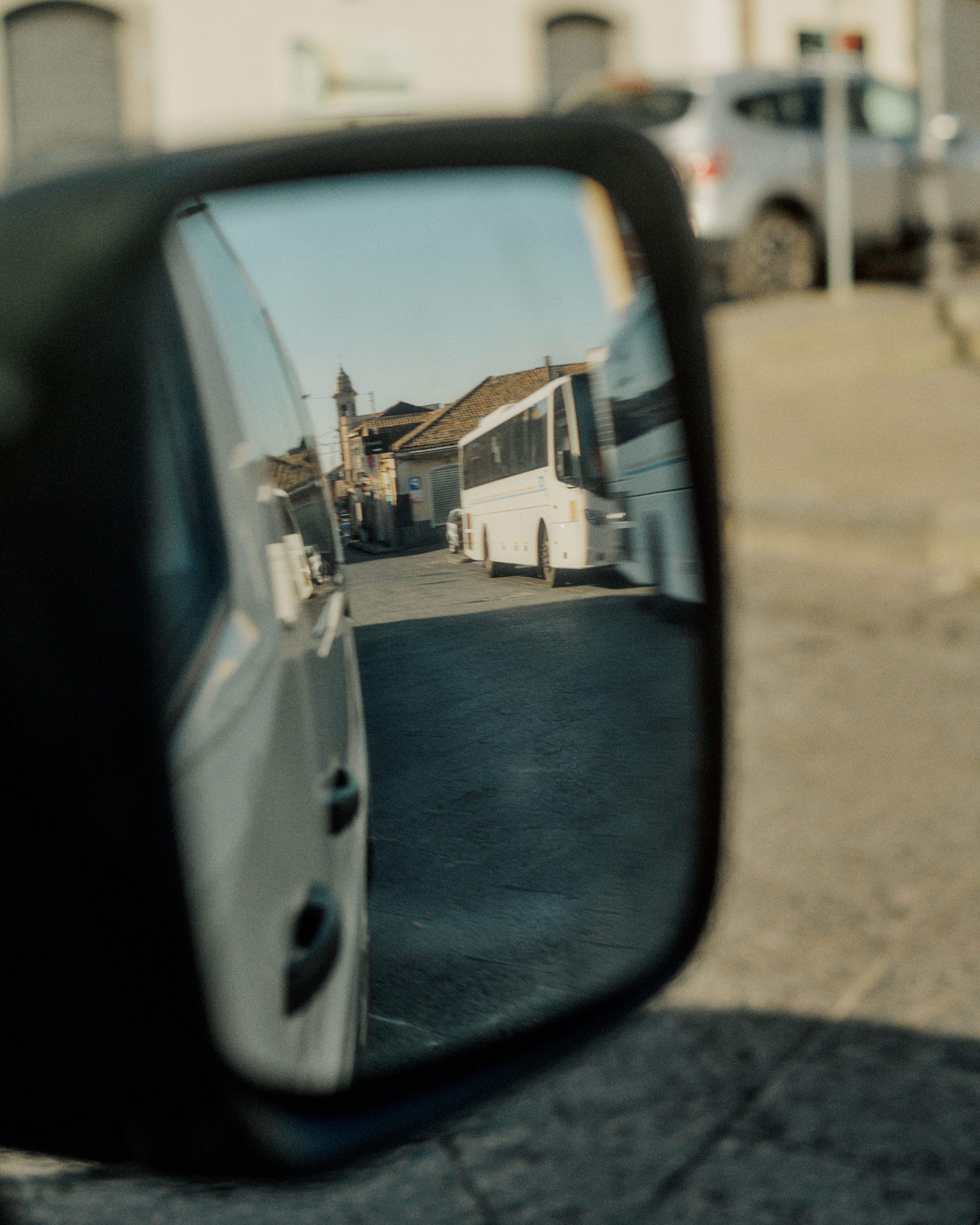 Car side mirror reflection showing Italian street scene with white bus and traditional buildings