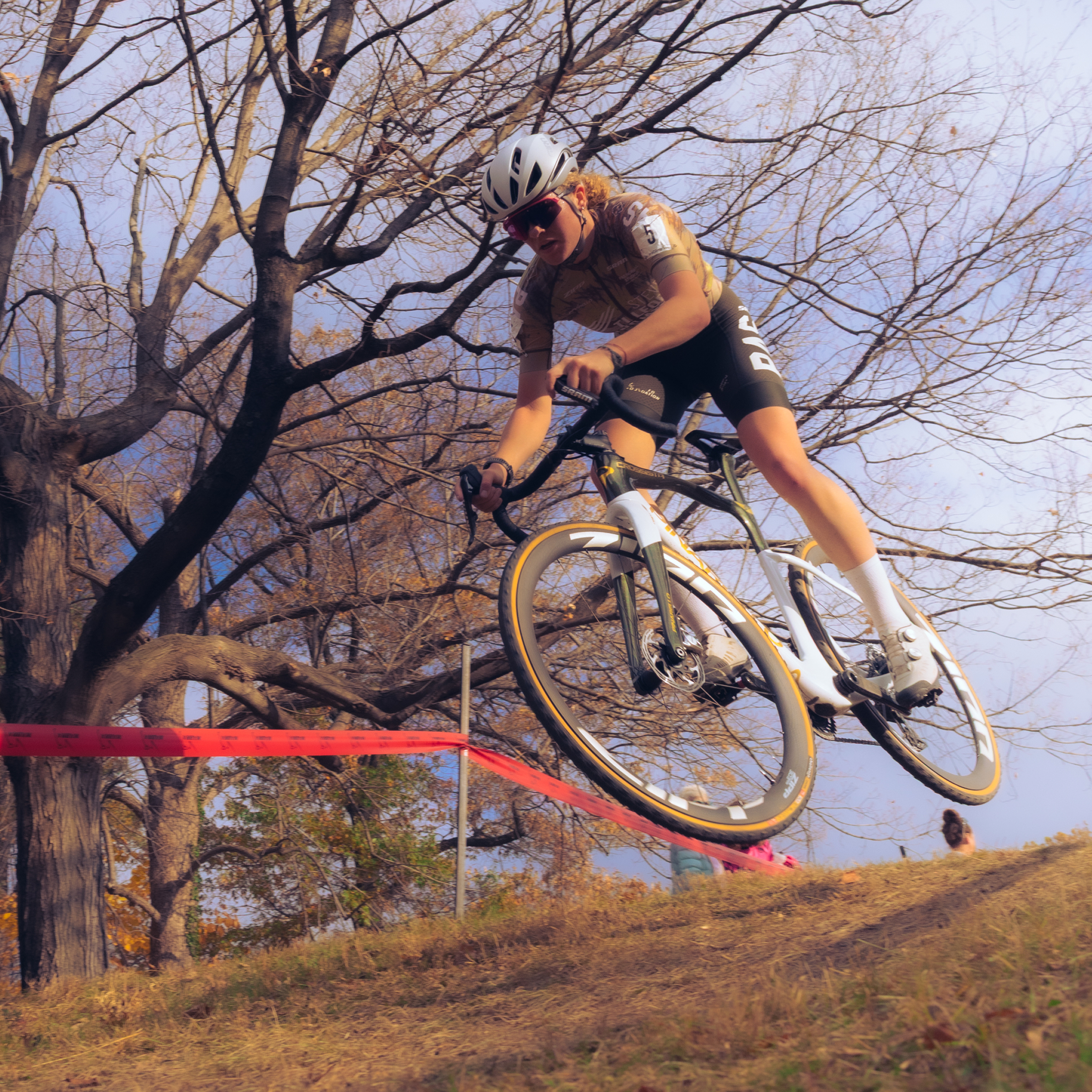 Model wearing white jersey jumping cyclocross bike over barrier during race