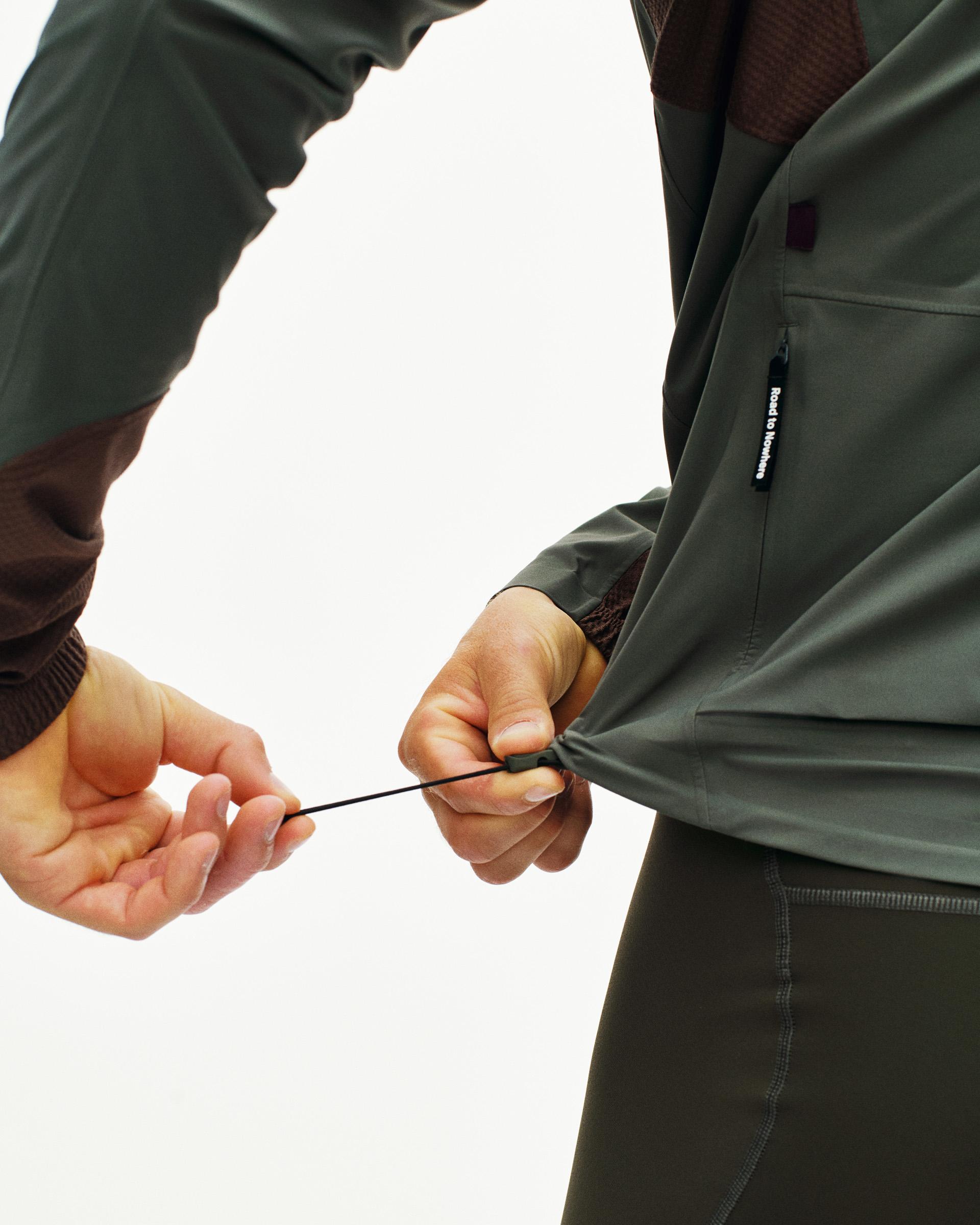 Dark Grey Mechanism Shell cycling jacket, detail on model showing drawcord hem adjustment with side zip pocket