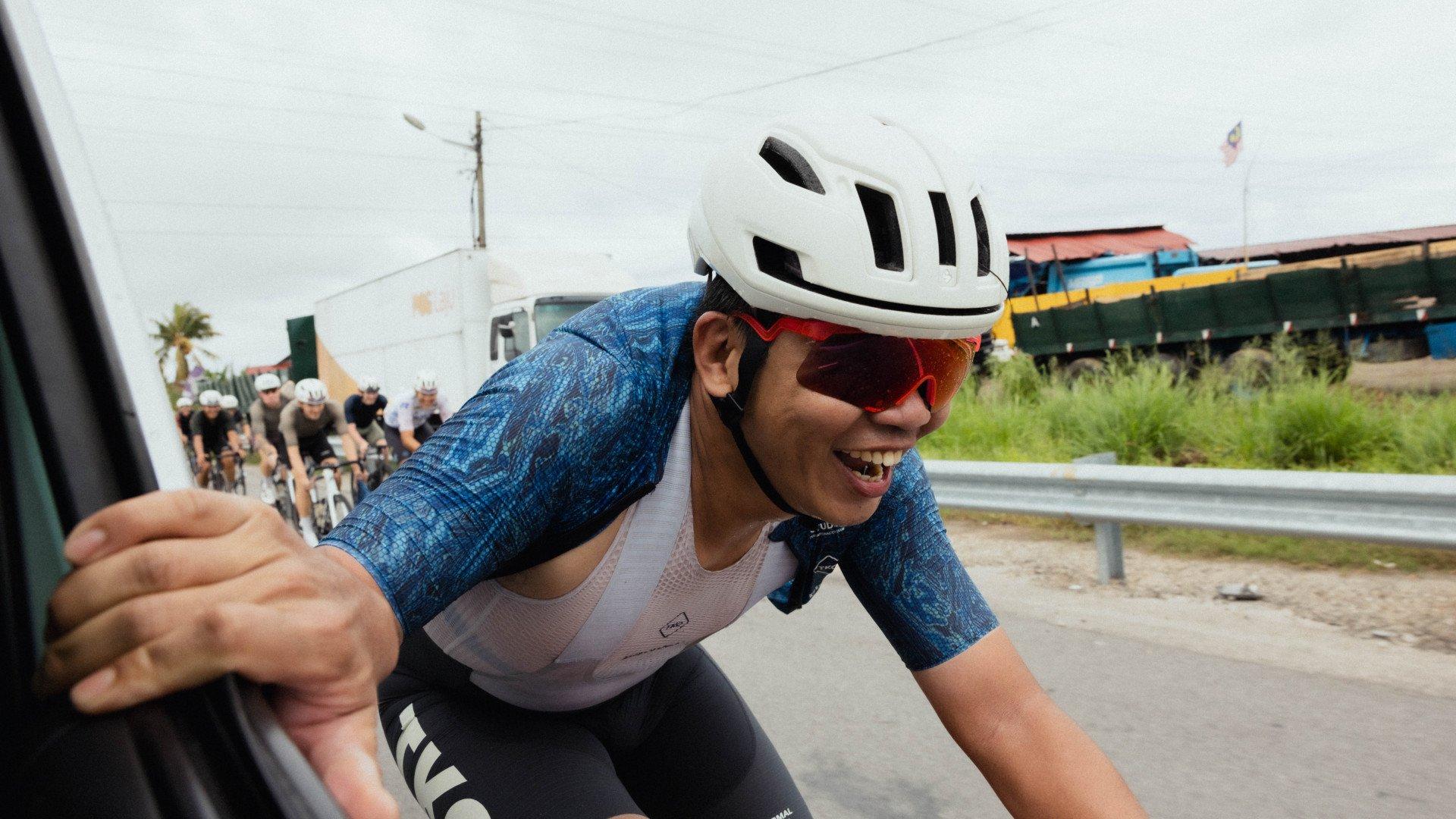 Model wearing blue patterned jersey and white base layer during cycling event, reaching toward camera with smile