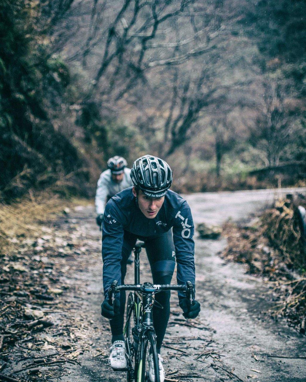 Two cyclists in dark training gear riding time trial bikes on forest road in aerodynamic position
