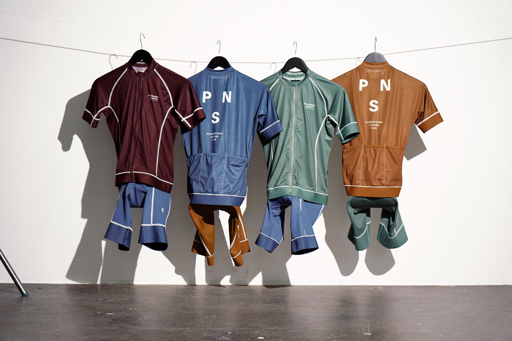 Four cycling jerseys and shorts sets hanging on clothesline in burgundy, blue, green, and orange colorways with logo
