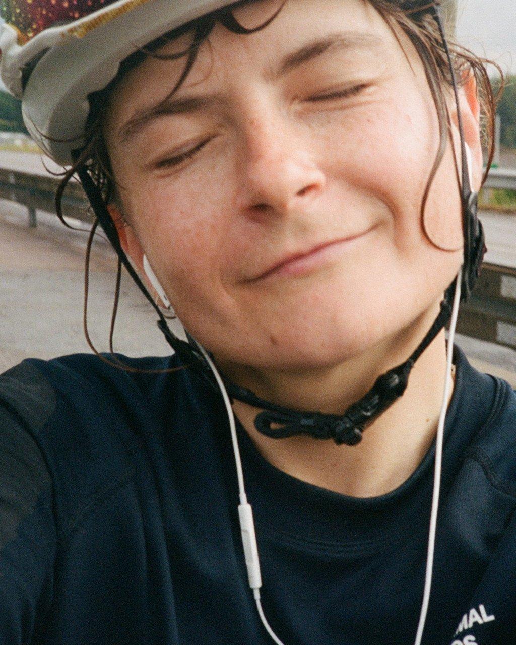 Model wearing navy blue jersey with white earphones, smiling with eyes closed while wearing cycling helmet