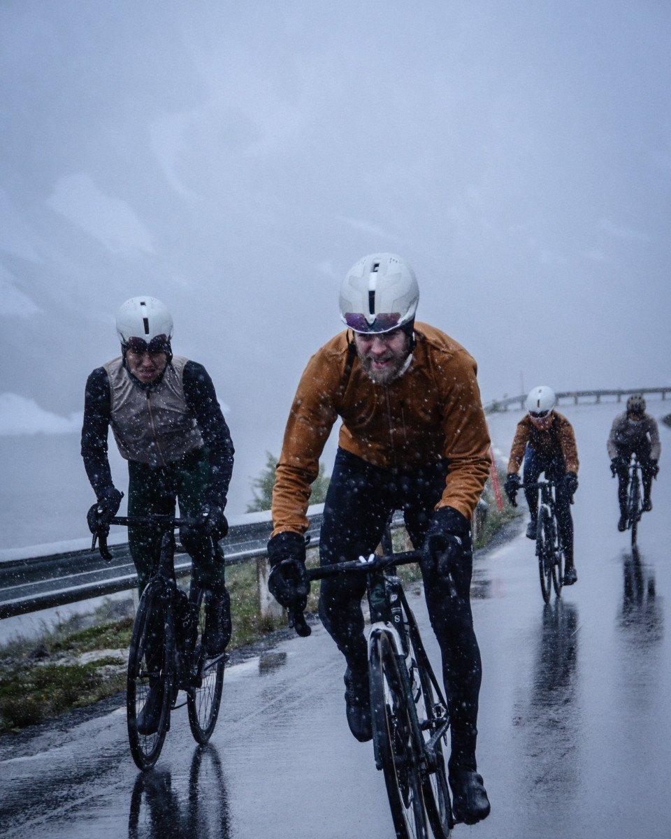 Cyclists riding in heavy snow on wet road wearing winter cycling gear including orange and dark jackets with helmets