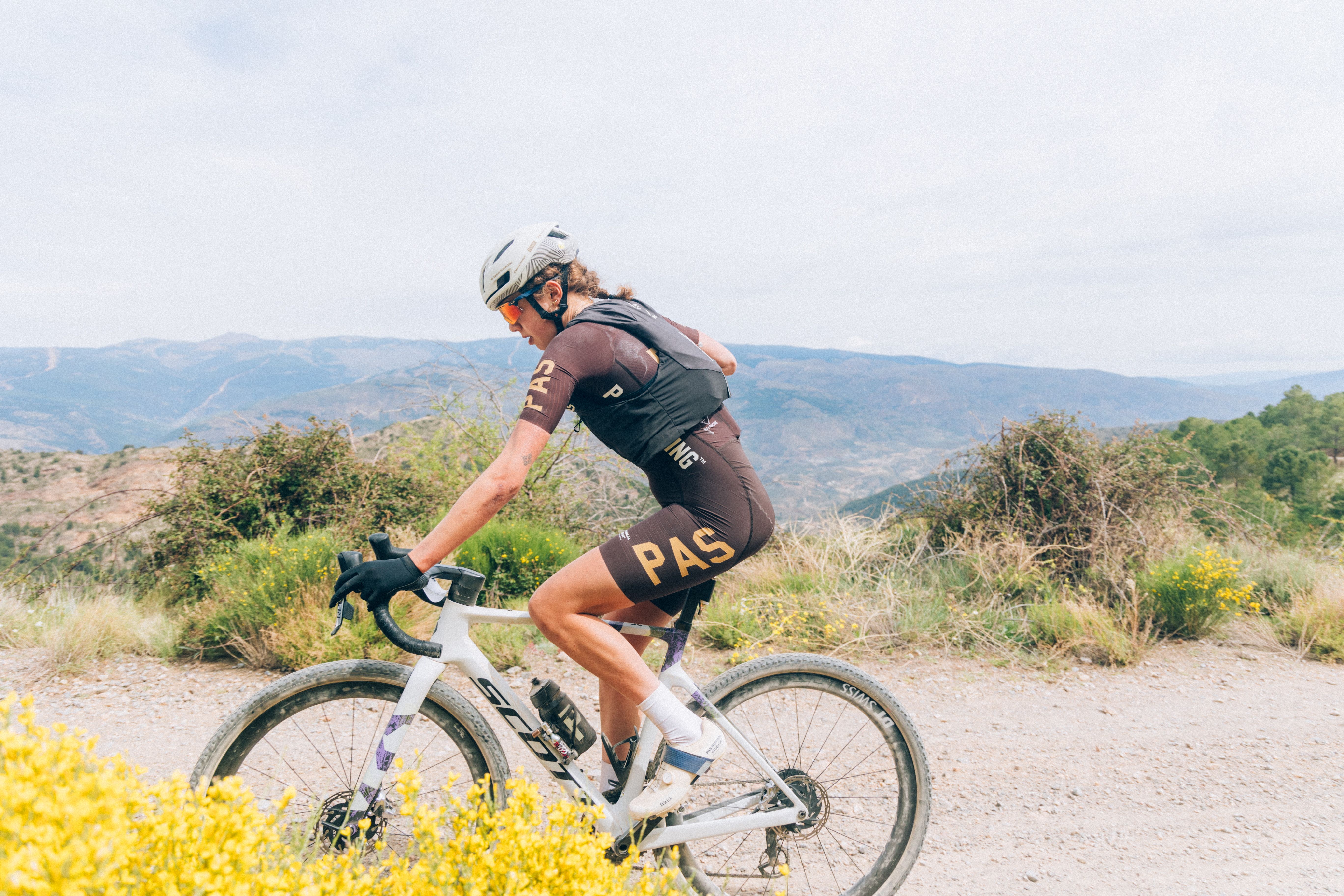 Model wearing brown PAS Racing cycling kit on gravel bike in mountainous terrain