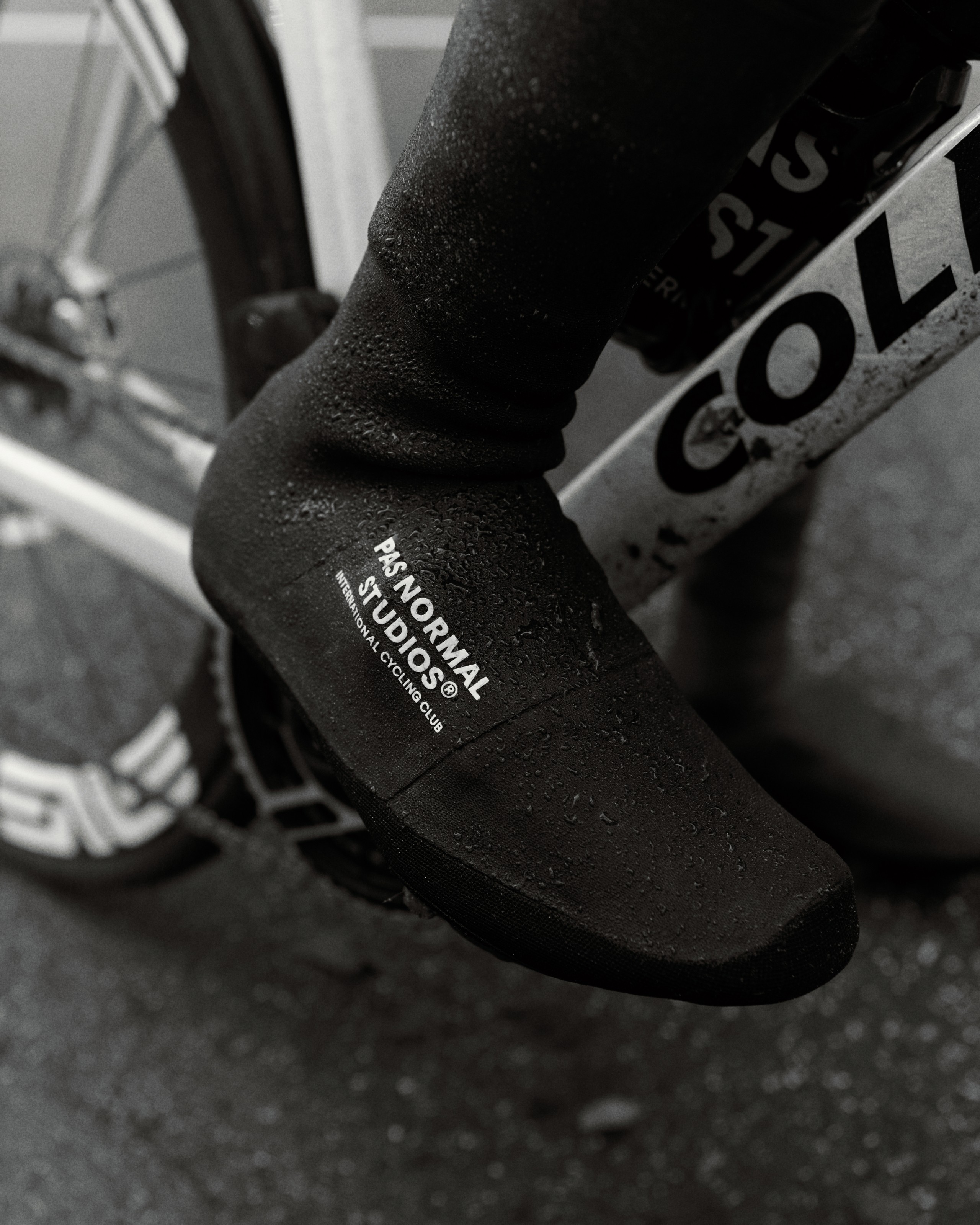 Black neoprene shoe cover with water droplets and brand text, providing thermal insulation and stretch fit
