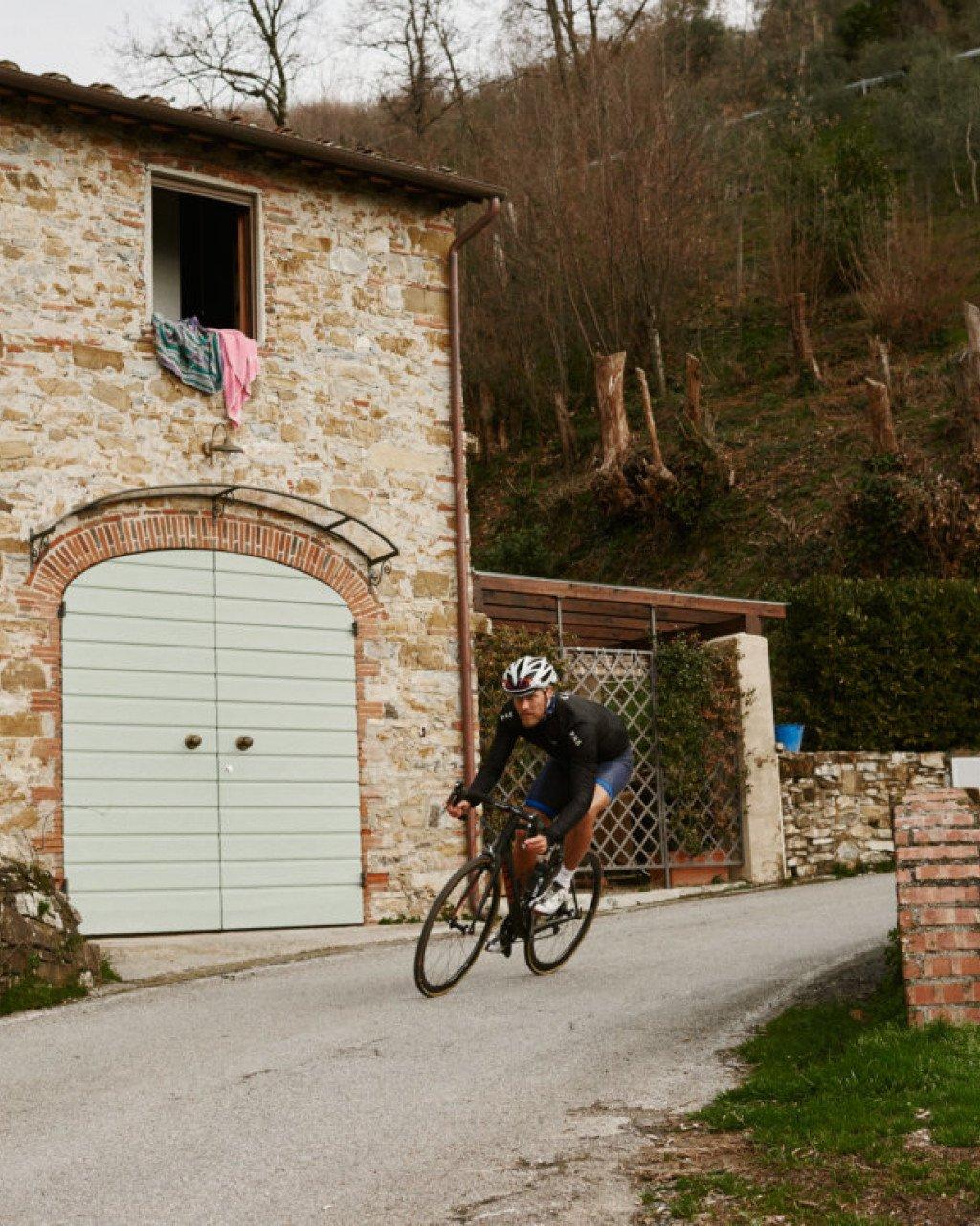 Model wearing black cycling jersey and blue shorts riding past stone building in Italian countryside