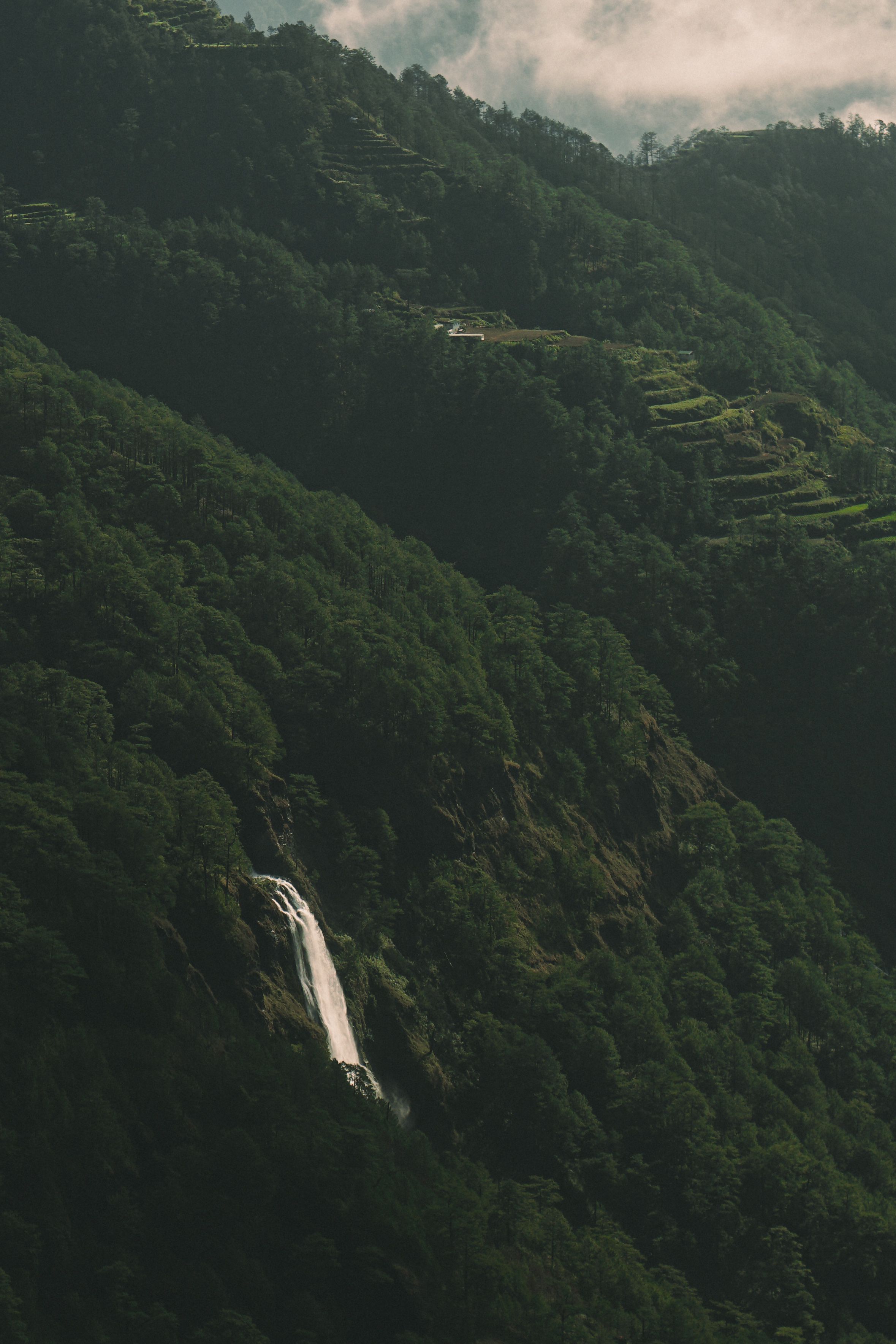 Lush green mountain valley with terraced hillsides and waterfall under cloudy sky