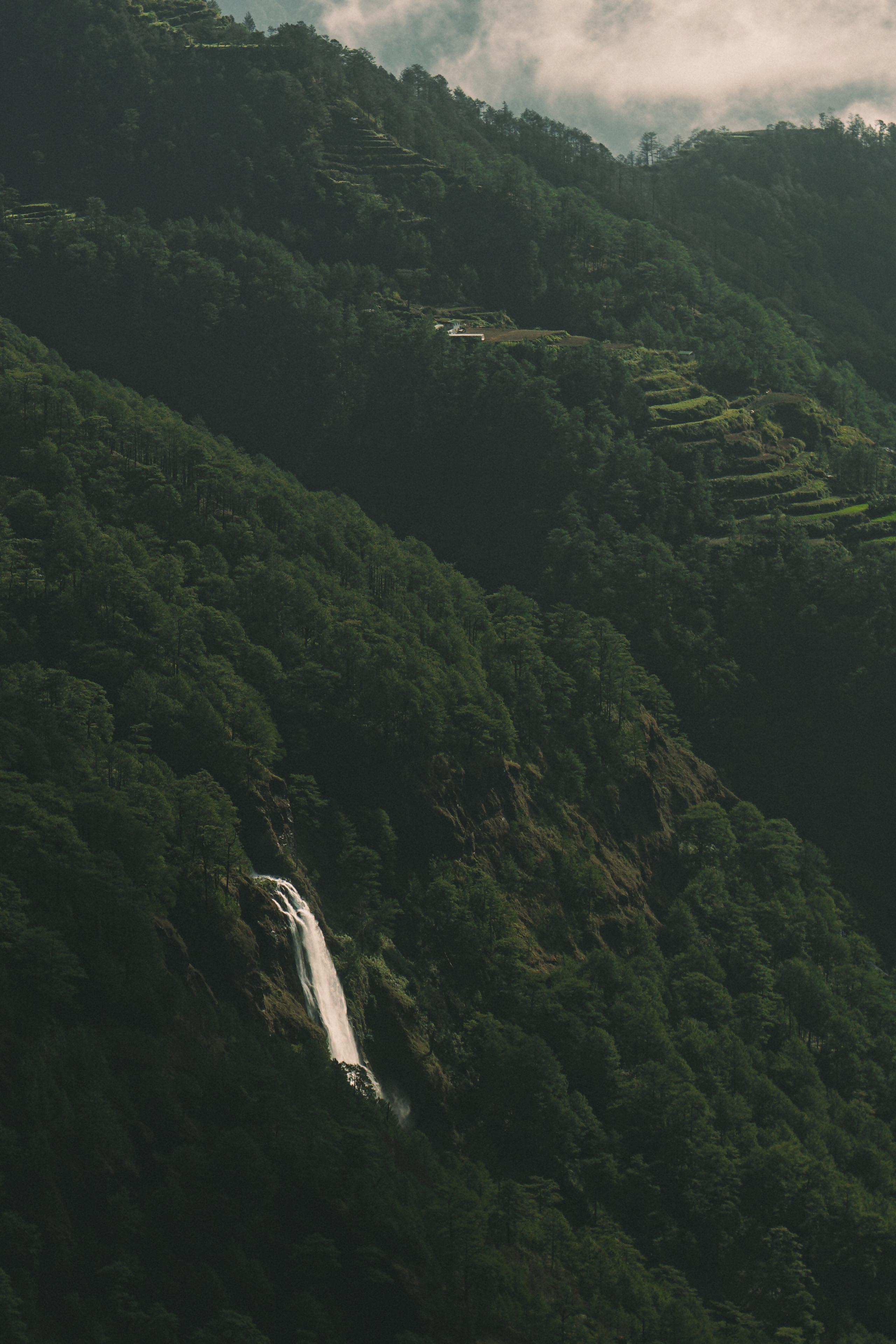Lush green mountain valley with terraced hillsides and waterfall under cloudy sky