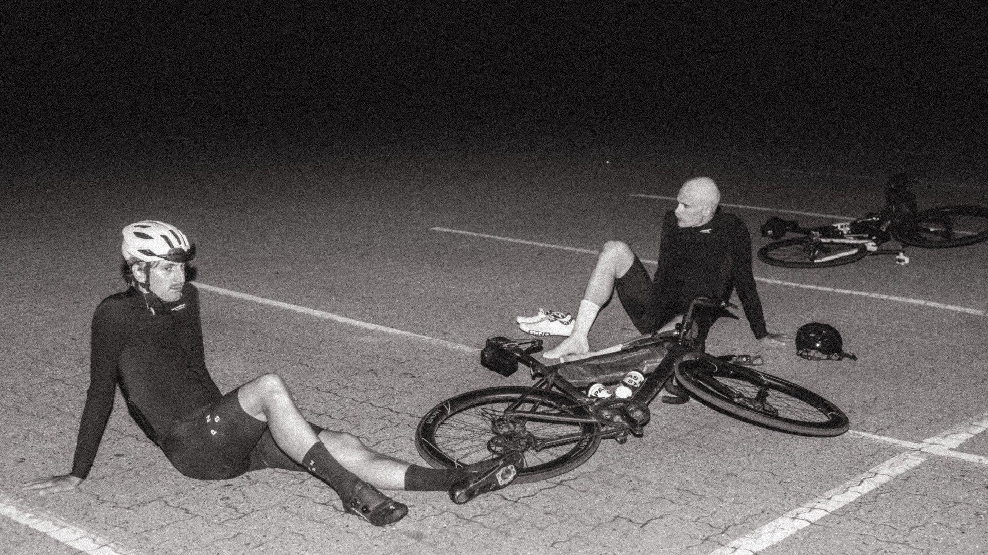 Two cyclists in black cycling apparel resting on road with bikes, black and white photography