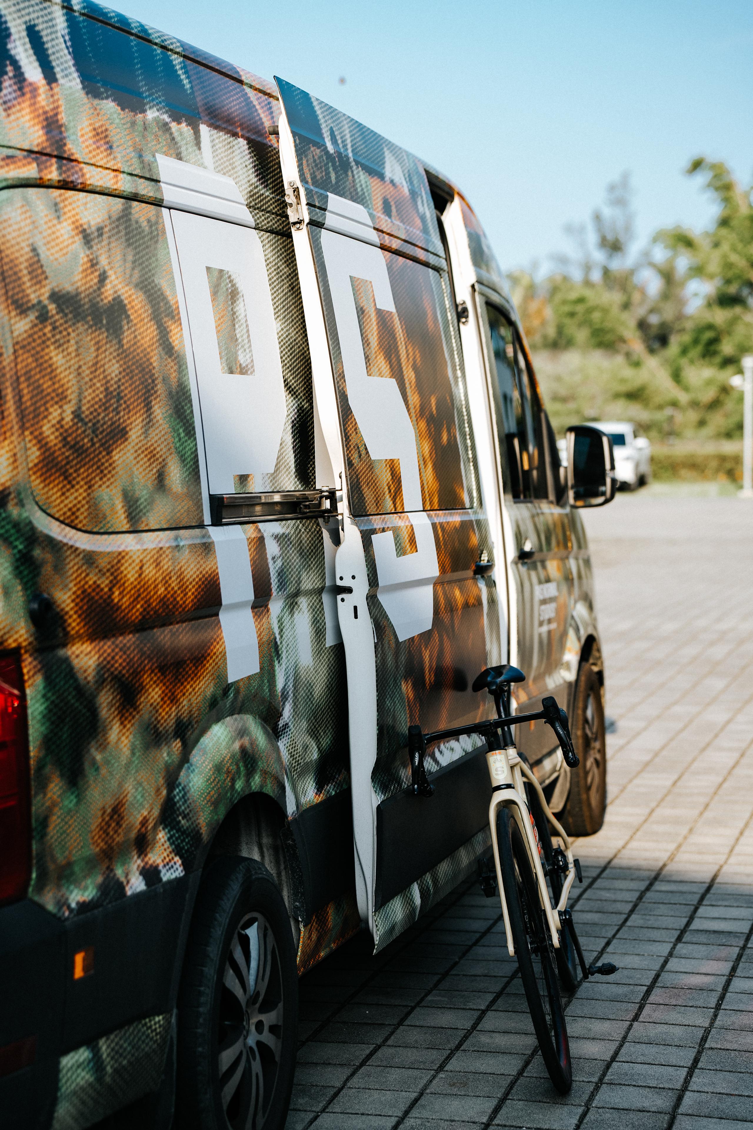 Team van with large logo branding and bicycle mounted on rear, parked on cobblestones