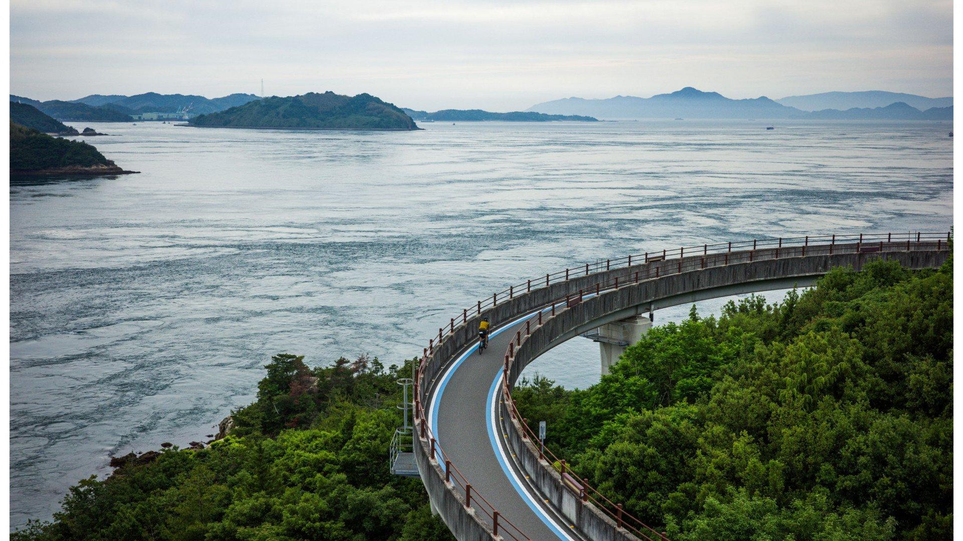 Cyclist on curved coastal bridge overlooking ocean with forested islands in Japan
