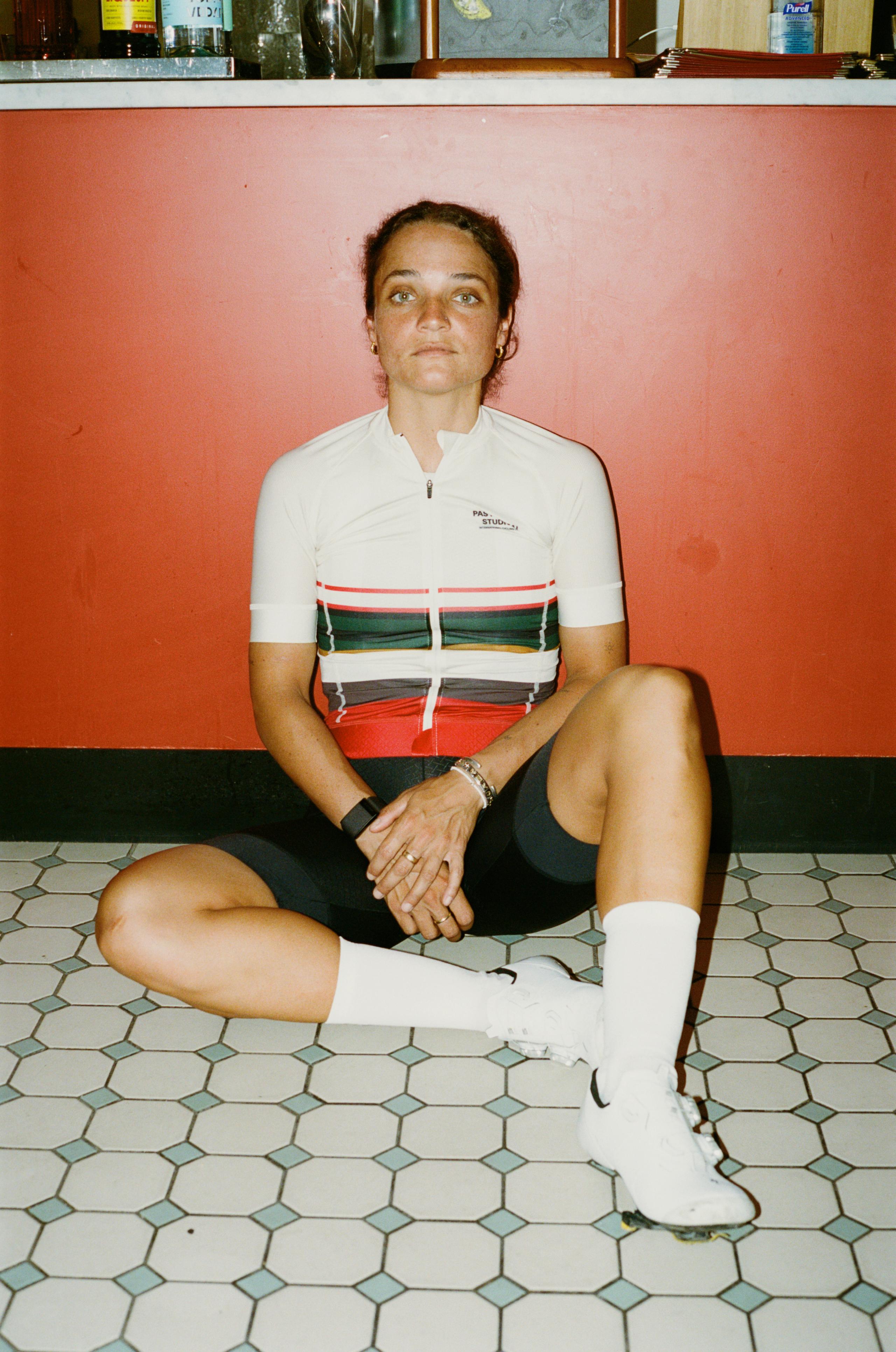 Model wearing white Mechanism jersey with red and green stripe pattern, sitting in casual pose